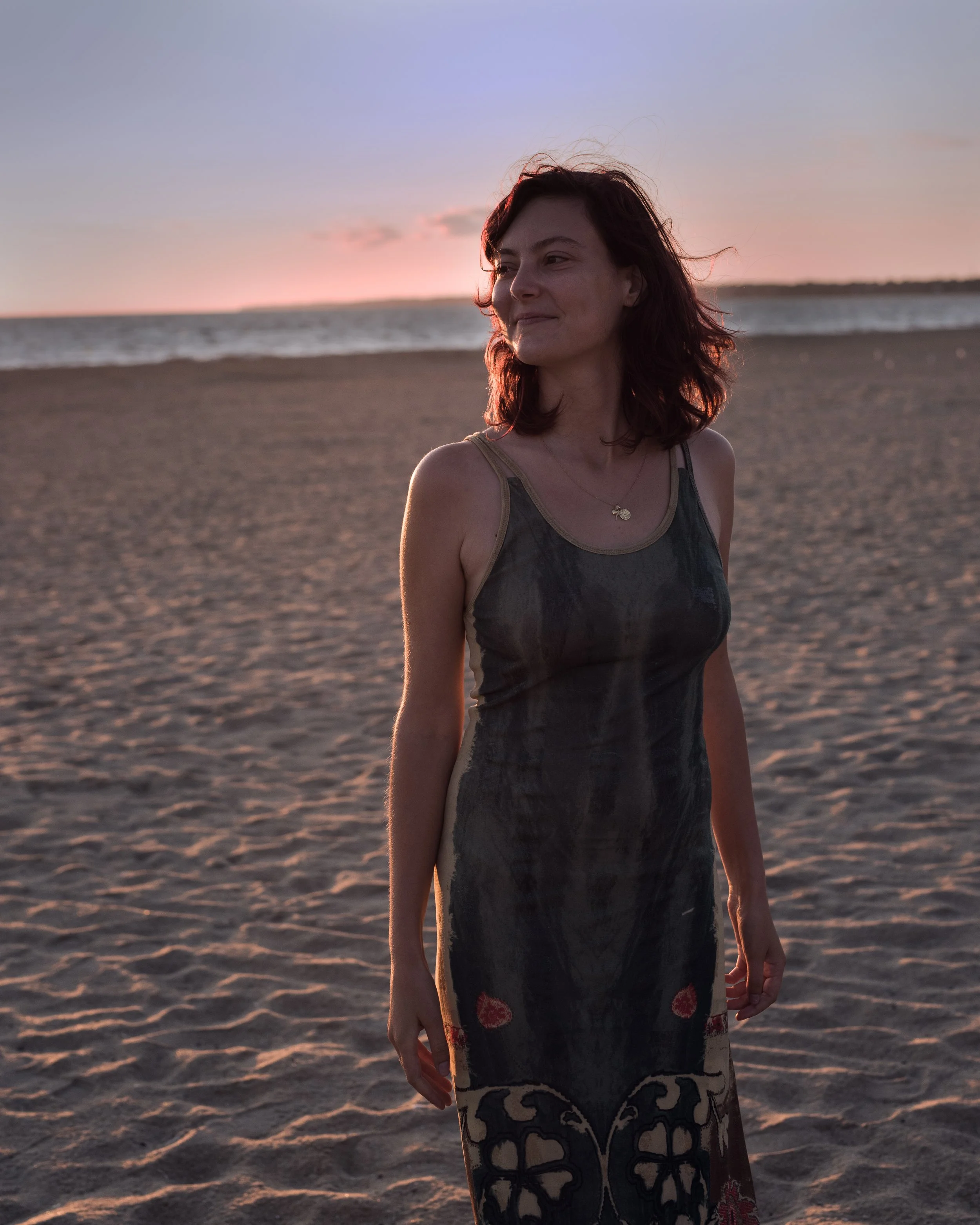 Femme debout sur la plage au coucher du soleil, regardant vers la droite, portant une robe noire et un collier, avec la mer en arrière-plan.