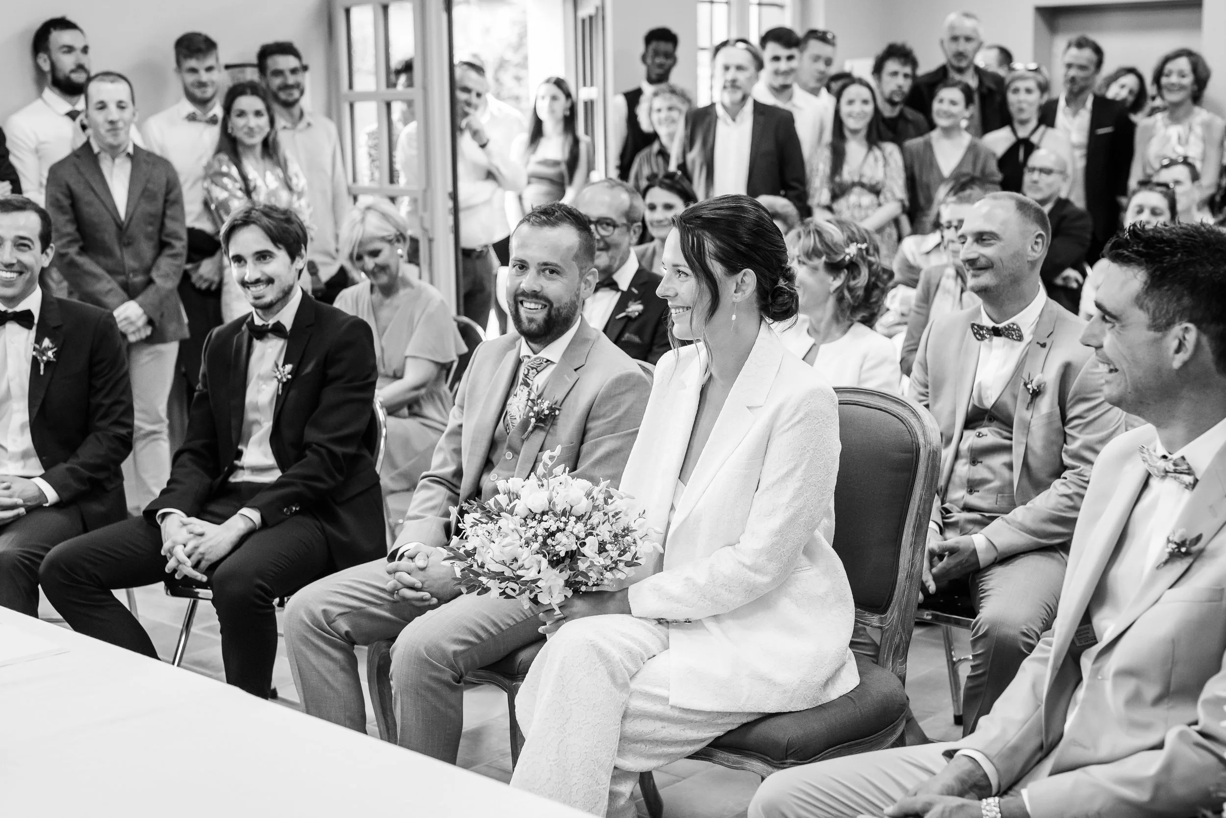 Groupe de personnes assises et debout lors d'une cérémonie, il s'agit probablement d'un mariage, avec des femmes en robe blanche et des hommes en costume.