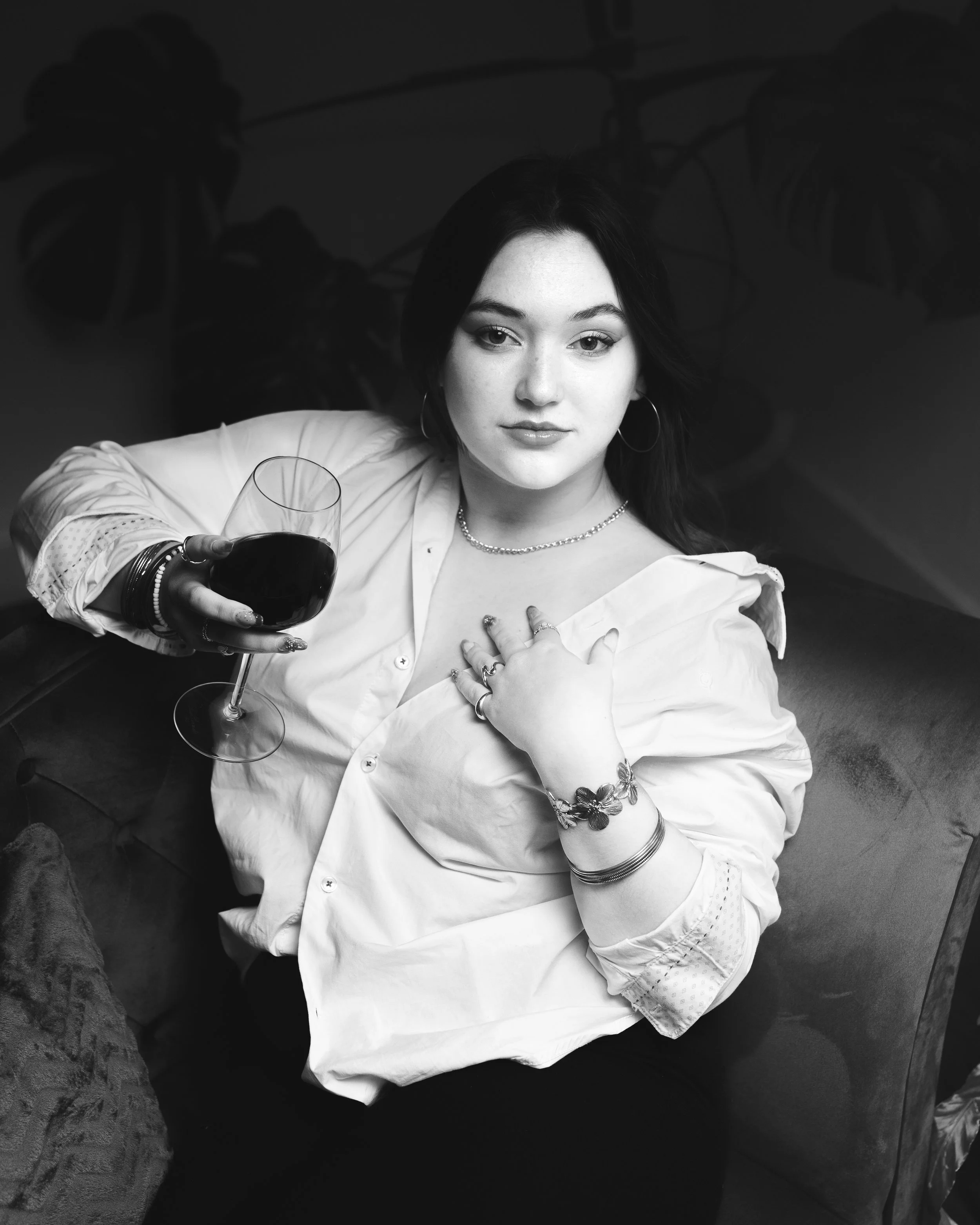 Une jeune femme élégante avec des boucles d'oreilles en anneaux, des bracelets et un collier, tient un verre de vin rouge et se pose la main sur la poitrine, avec un fond de plantes en grande feuille.
