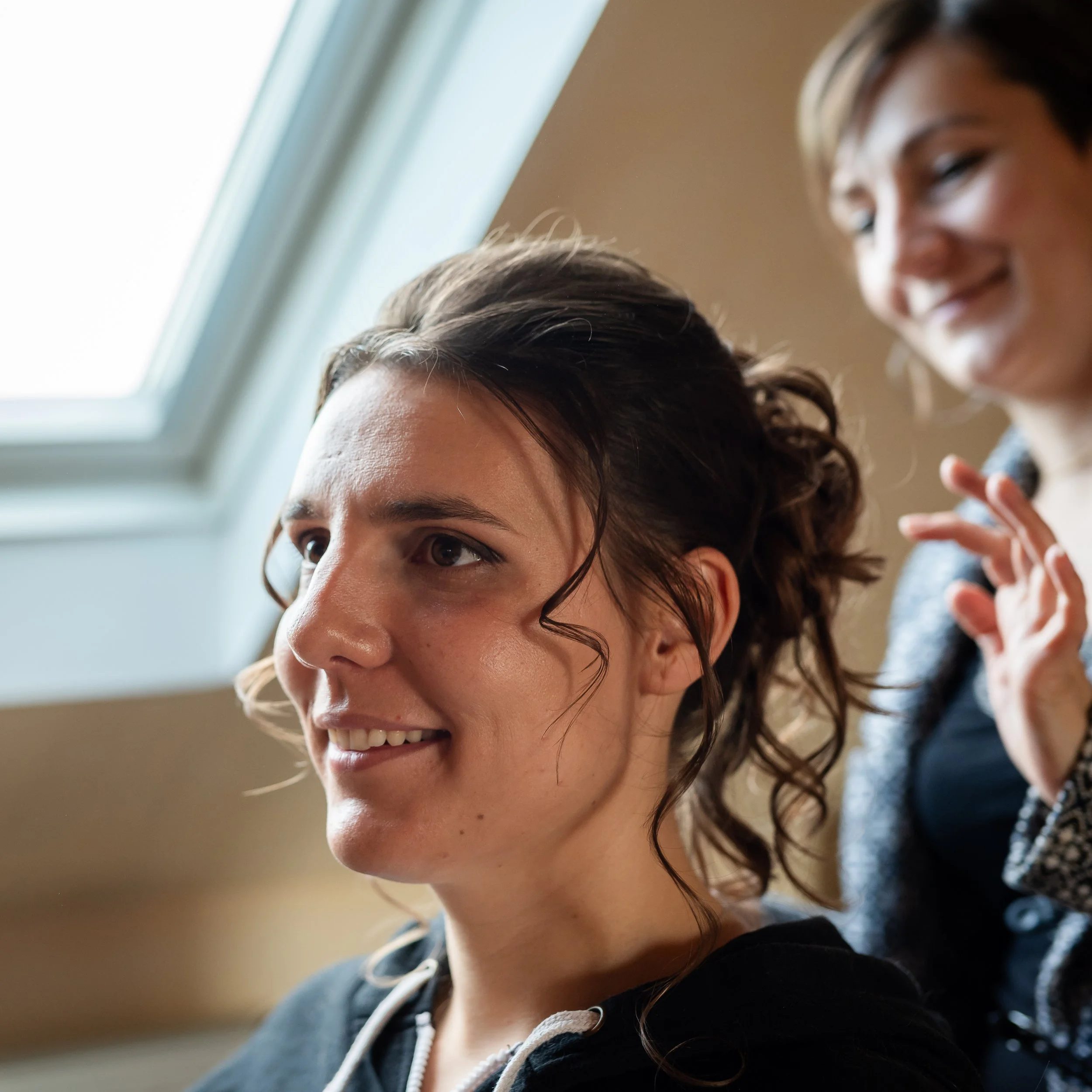 Premier plan d'une jeune femme souriante avec des cheveux bruns bouclés, portant un vêtement noir avec une fermeture éclair, dans un intérieur avec une fenêtre à l'arrière.