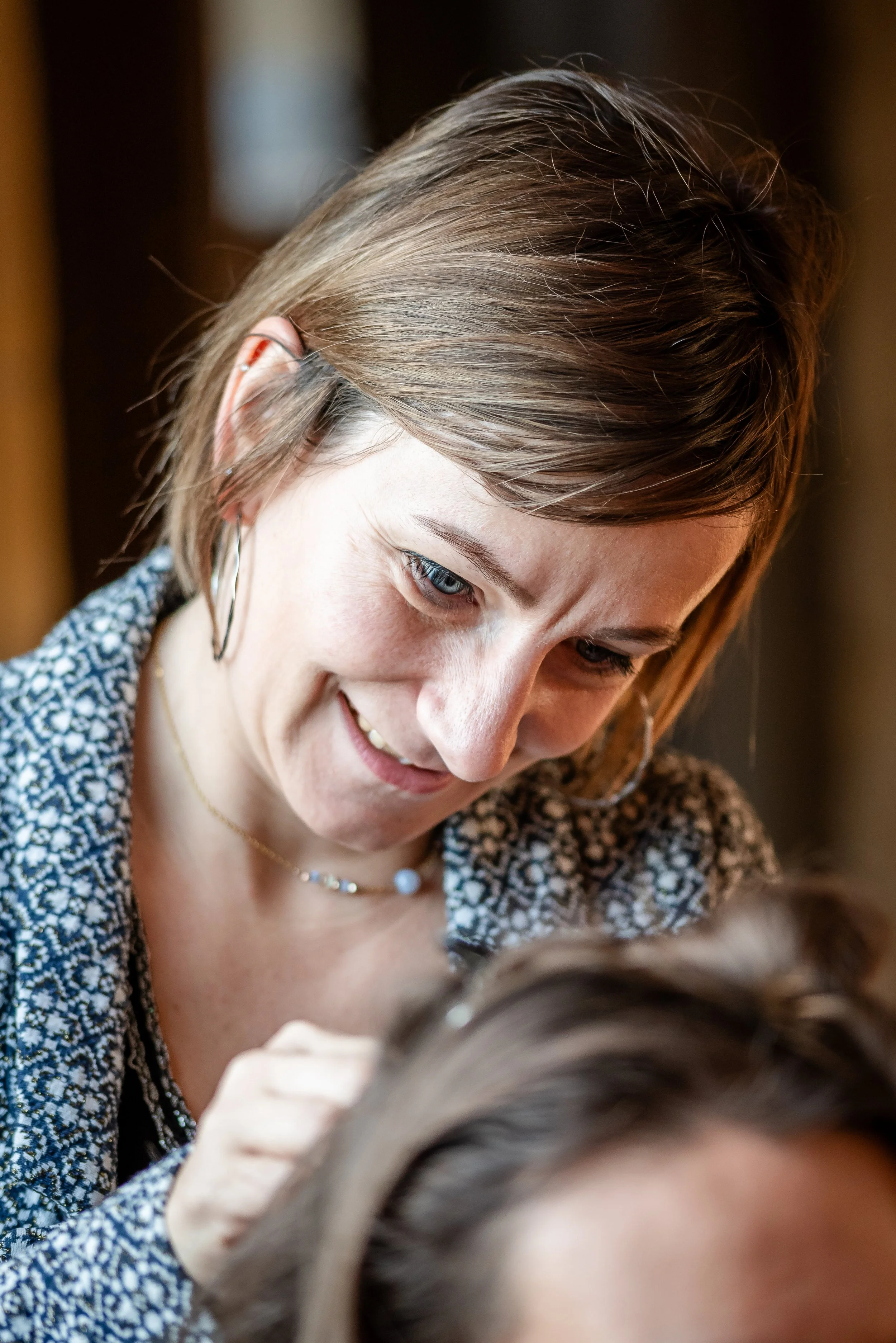 Une femme souriante avec des cheveux courts, portant une robe à motifs et un collier, qui regarde vers un enfant dont on ne voit que les cheveux.