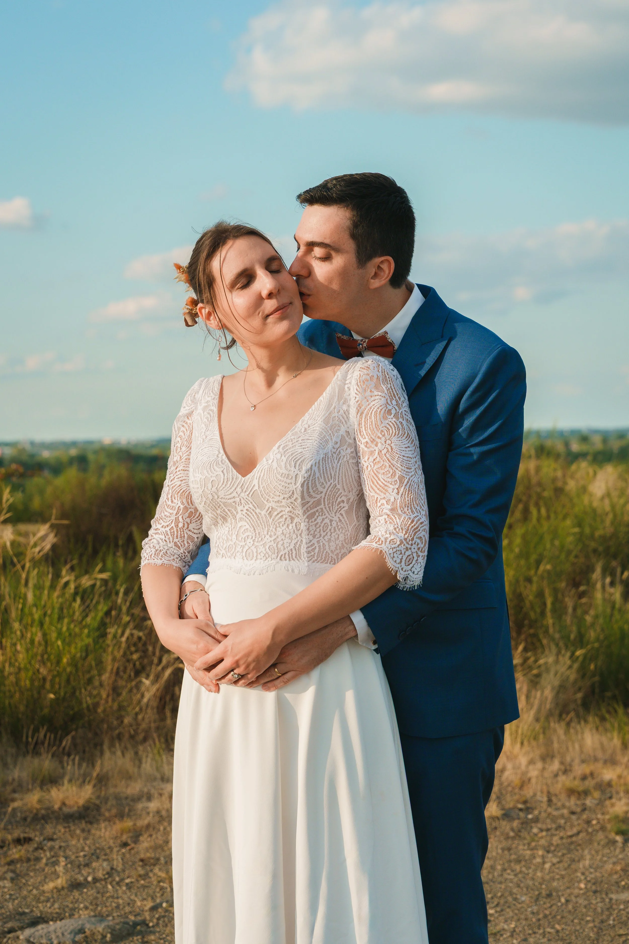 Un couple en tenue de mariage dans la nature, l'homme embrassant la femme sur la joue, ciel bleu avec quelques nuages à l'arrière-plan.