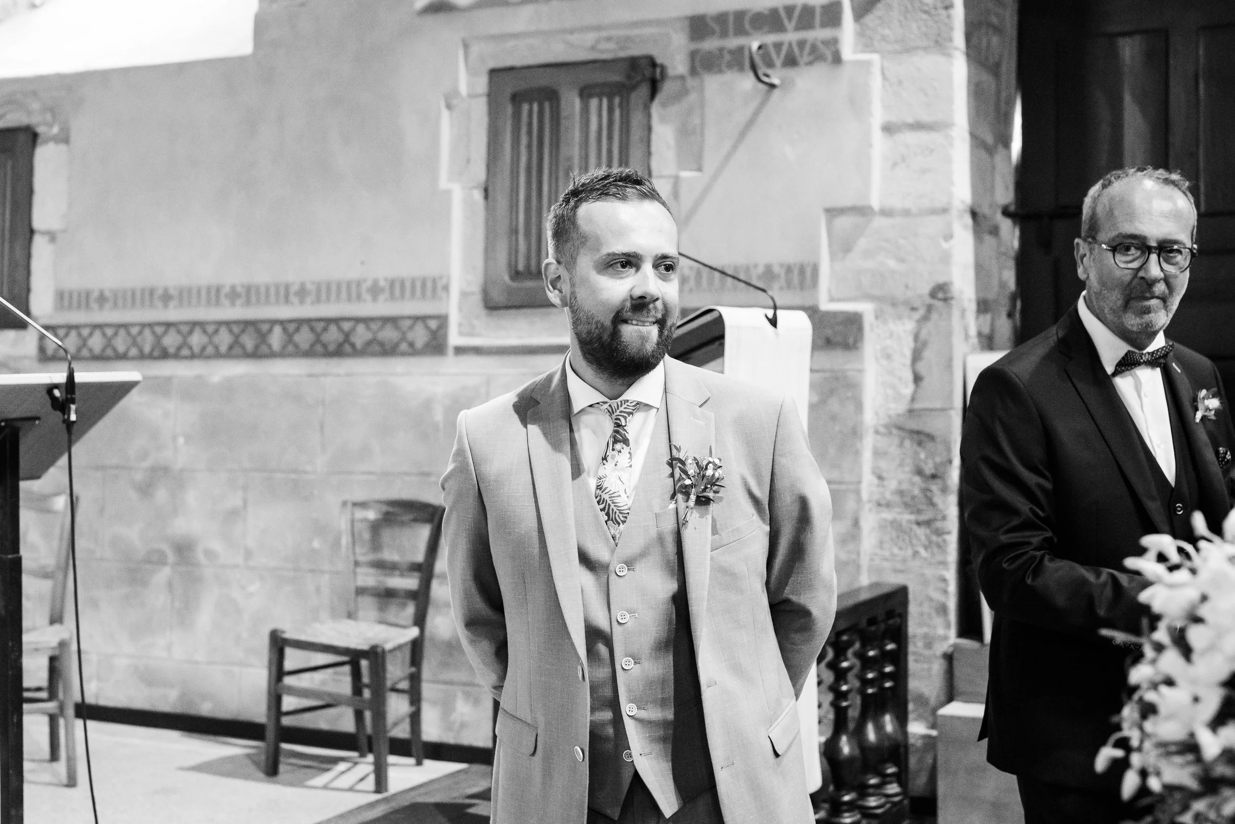 Deux hommes en costume lors d'une cérémonie, l'un souriant avec une boutonnière, l'autre portant des lunettes, dans une salle aux murs en pierre.