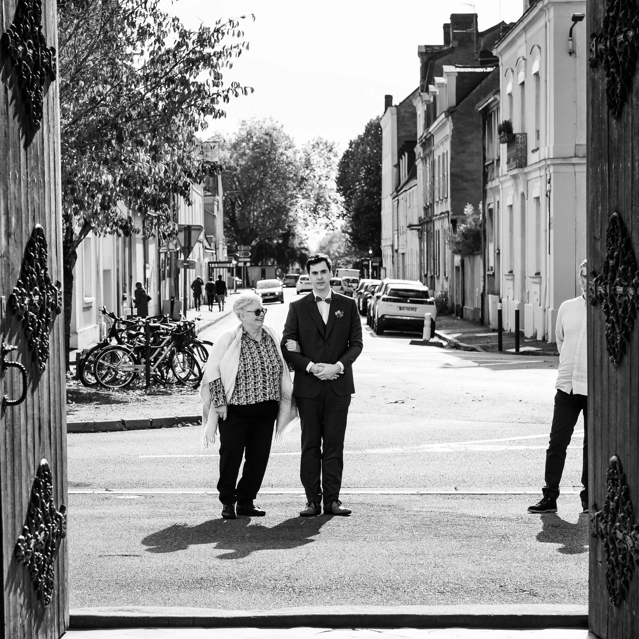 Une entrée d'un jardin ou d'une propriété, avec deux portes en acier ouvrant sur une rue pavée. De part et d'autre, il y a des arbres et des bâtiments résidentiels. Au premier plan, deux personnes, une femme âgée et un homme jeune en costume, marchen