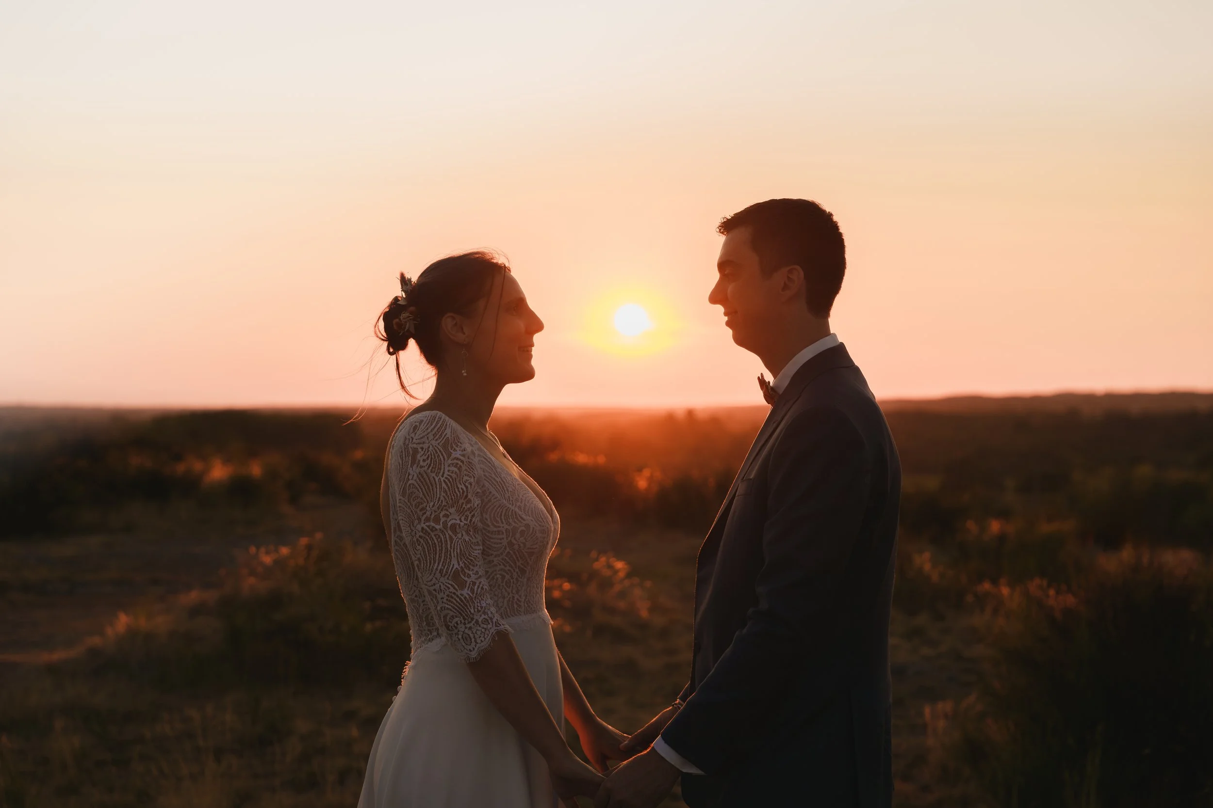 Un couple en costume de mariage se tenant la main face à face lors d'un coucher de soleil en plein air.