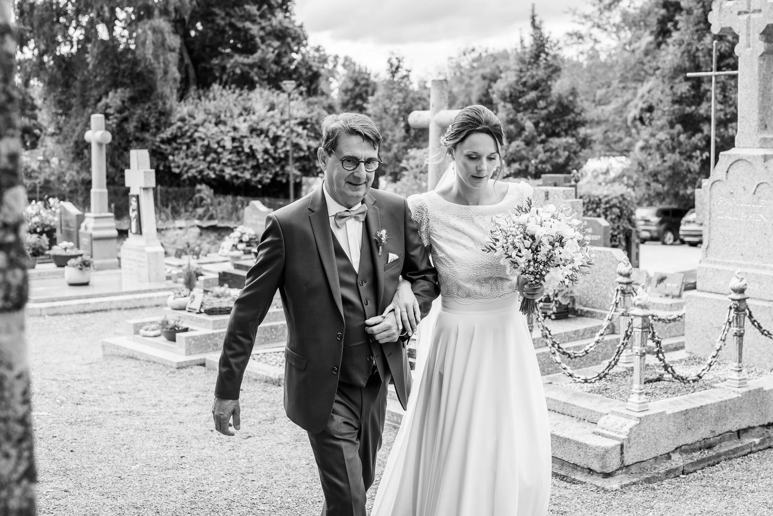 Un couple de mariés marche dans un cimetière, tenant la main. La femme porte une robe de mariage et tient un bouquet de fleurs, tandis que l'homme porte un costume sombre avec une chemise blanche et une cravate ou un nœud papillon.
