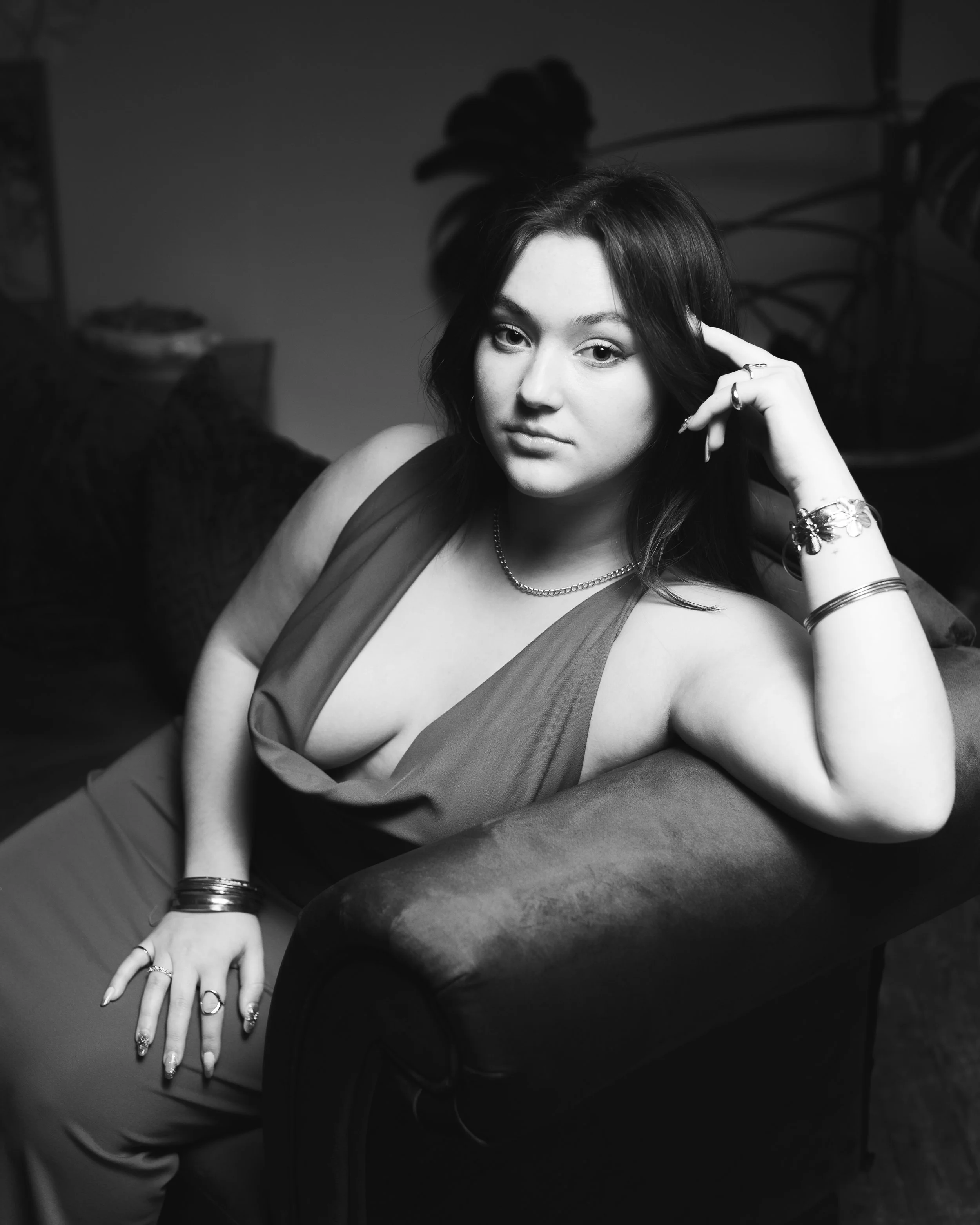 Une femme pose sur un canapé en noir et blanc, portant une robe à baillettes, avec des bijoux et plusieurs bagues. Elle a les cheveux longs et foncés, et regarde l'appareil photo.