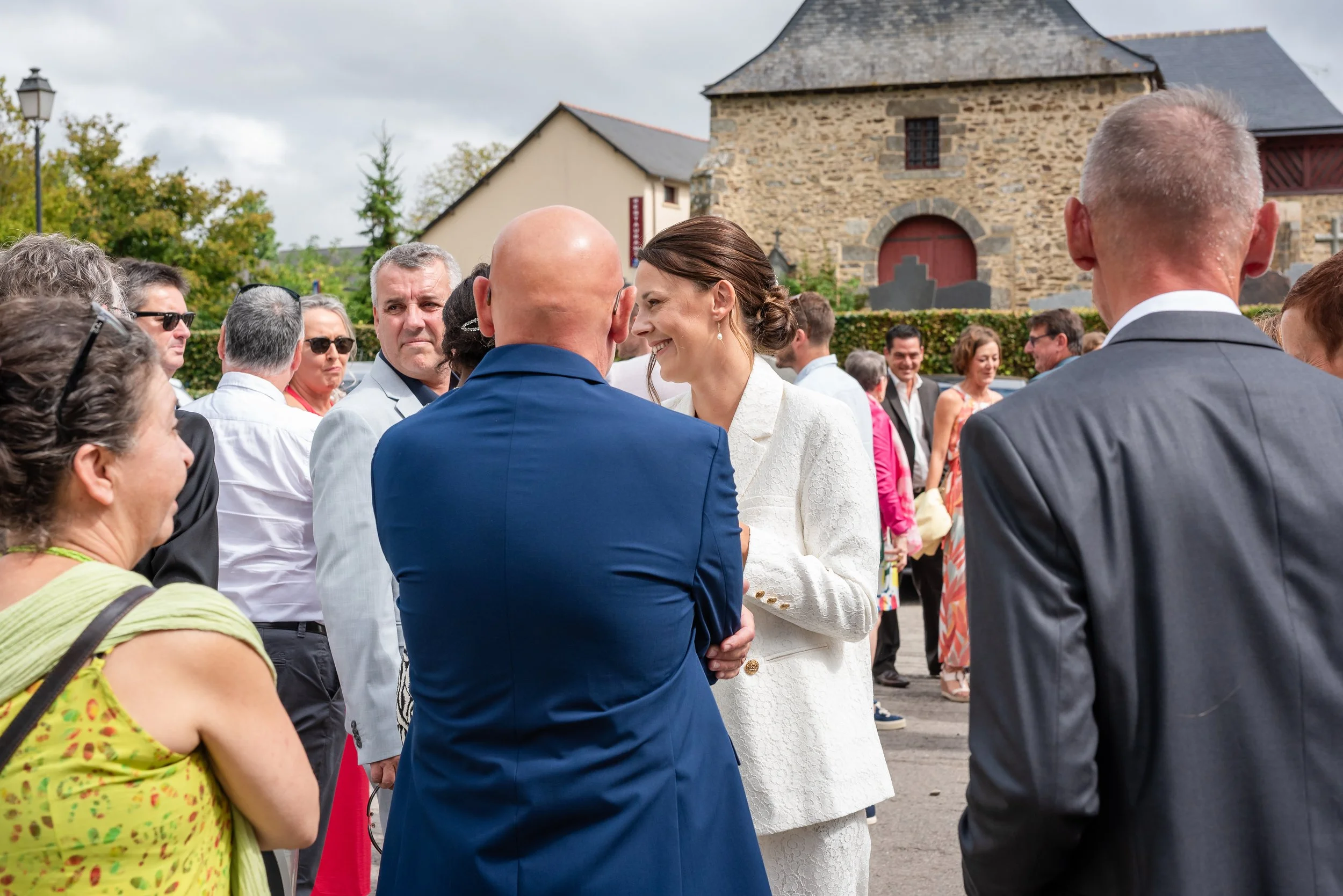 Un groupe de personnes, dont un couple au centre de la photo, échange lors d'une cérémonie extérieure devant un bâtiment en pierre. La femme porte une robe blanche et l'homme un costume bleu.