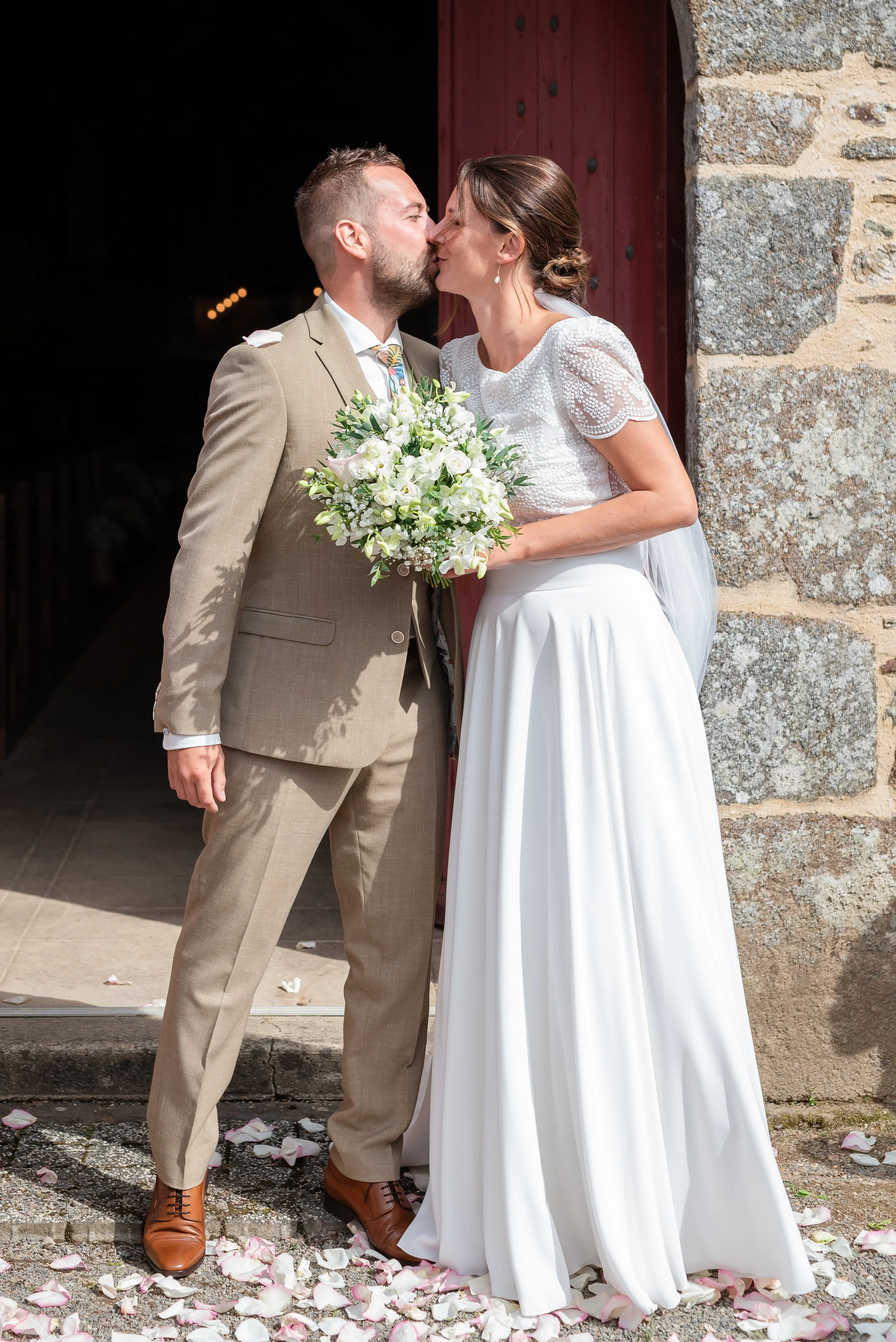 Un couple en mariage s'embrasse devant une porte en pierre, entouré de pétales de roses.