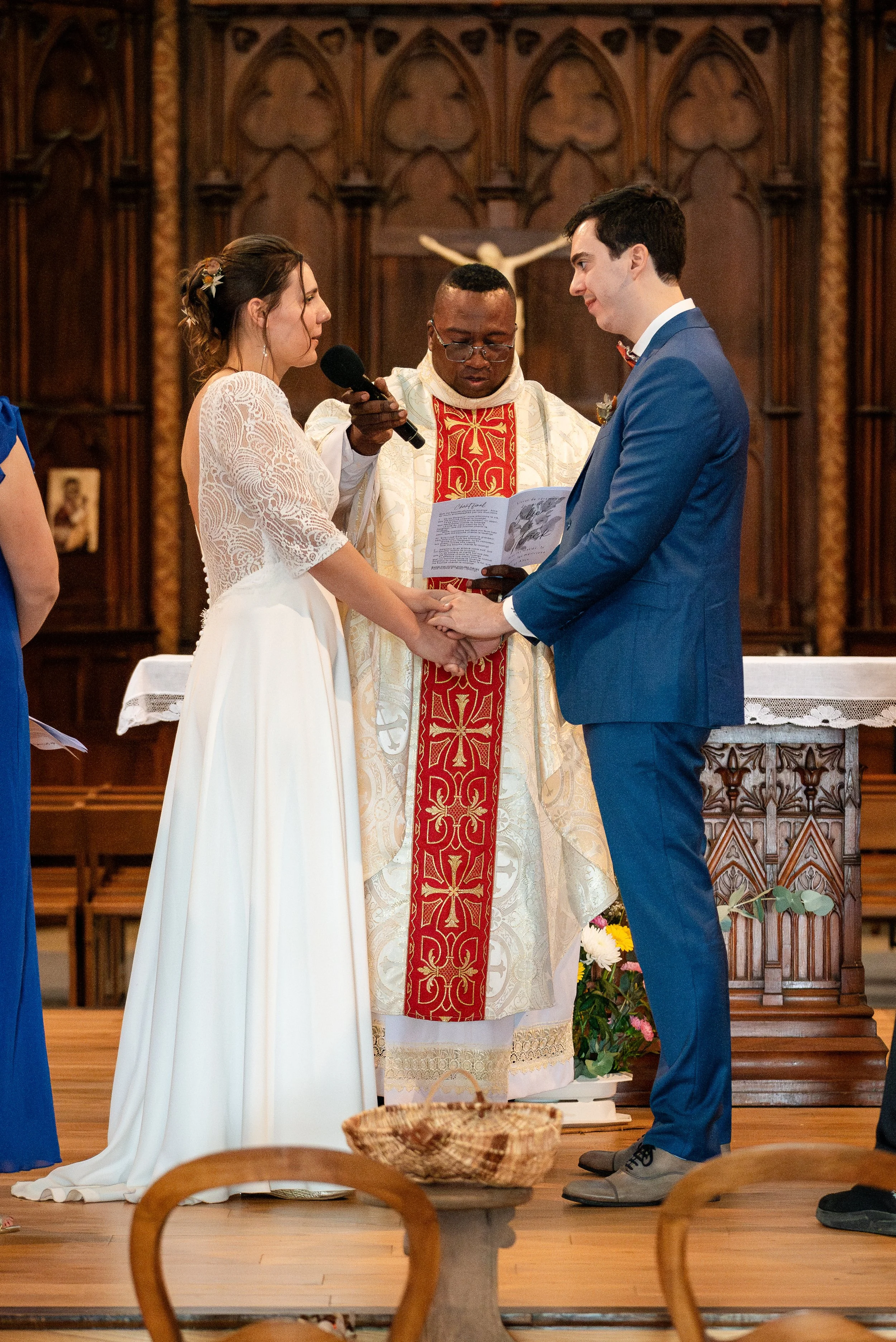 Un couple échange des vœux lors d'une cérémonie de mariage dans une église en bois. La mariée porte une robe blanche en dentelle, le marié un costume bleu. Un prêtre en robe cérémoniale anime la cérémonie, et le couple se tient la main, regardant dan