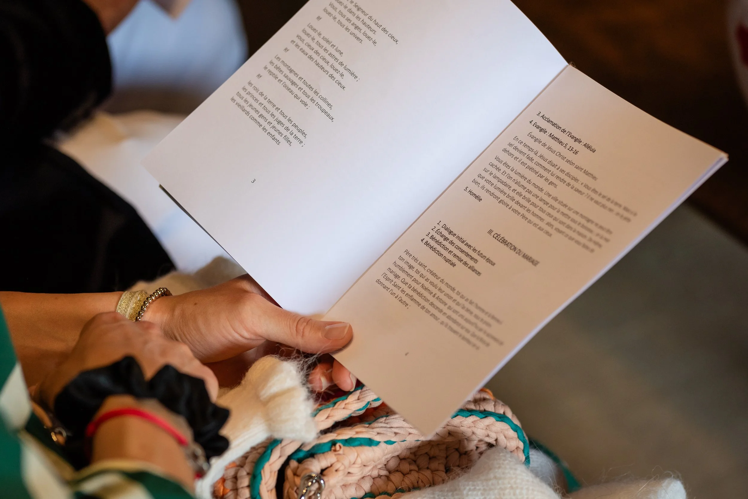 Mains d'une personne tenant un livre ouvert, portant plusieurs bracelets colorés. Fond flou avec des éléments tricotés et des vêtements.