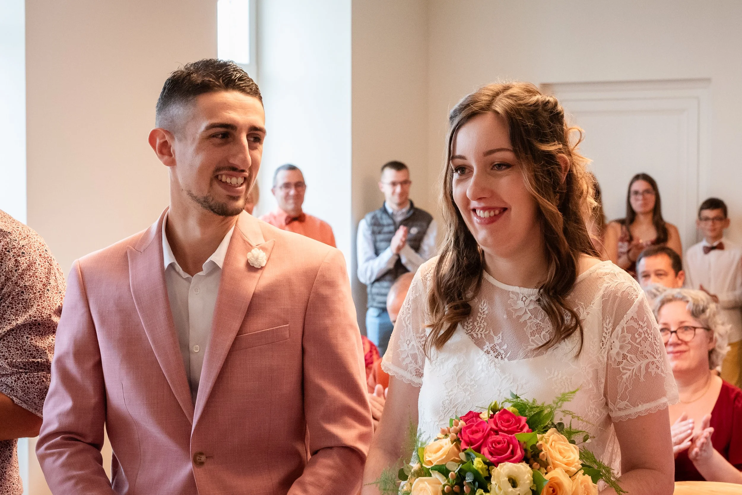Un couple lors d'une cérémonie de mariage. La femme porte une robe en dentelle blanche et tient un bouquet de fleurs roses, oranges et blanches. L'homme porte un costume rose pâle avec une chemise blanche. Ils sourient et regardent devant eux, entour