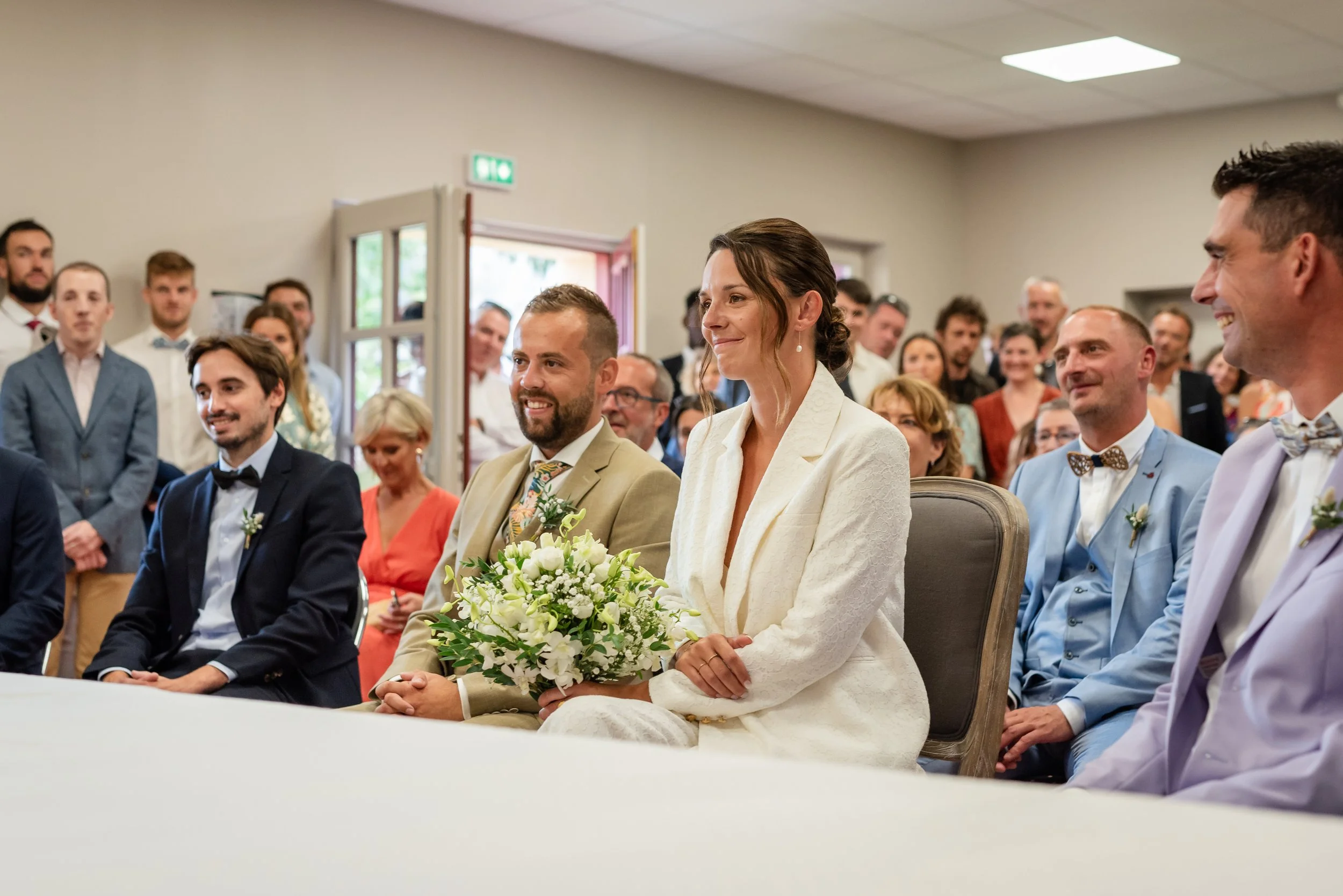 Un groupe de personnes assises lors d'une cérémonie, probablement un mariage, avec la mariée au centre tenant un bouquet de fleurs avec un blazer beige et une robe blanche