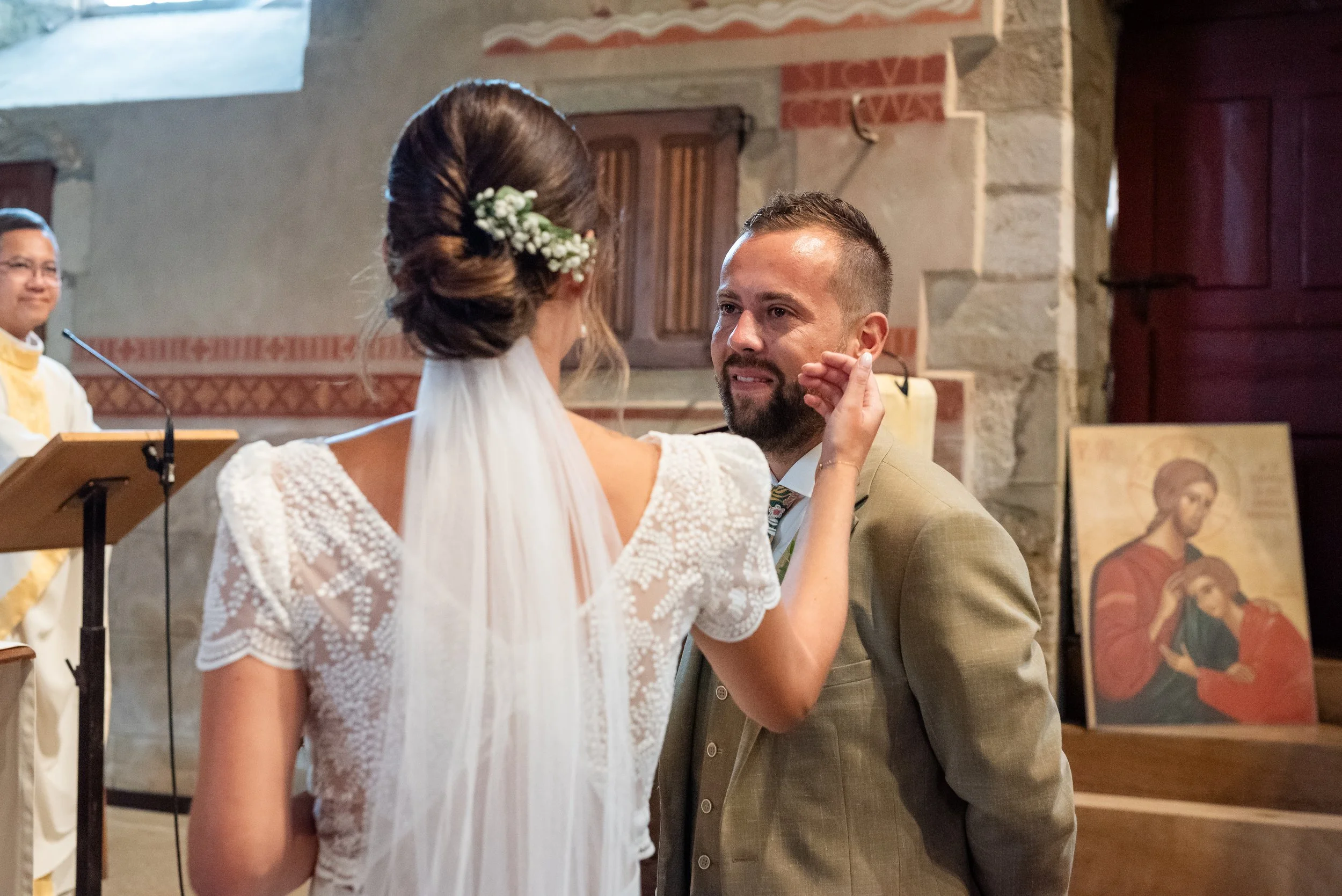 Un mariage à l'église avec un couple échangeant un acte, avec une femme en robe blanche à l'avant et un homme en costume beige, entourés d'autres personnes lors de la cérémonie.