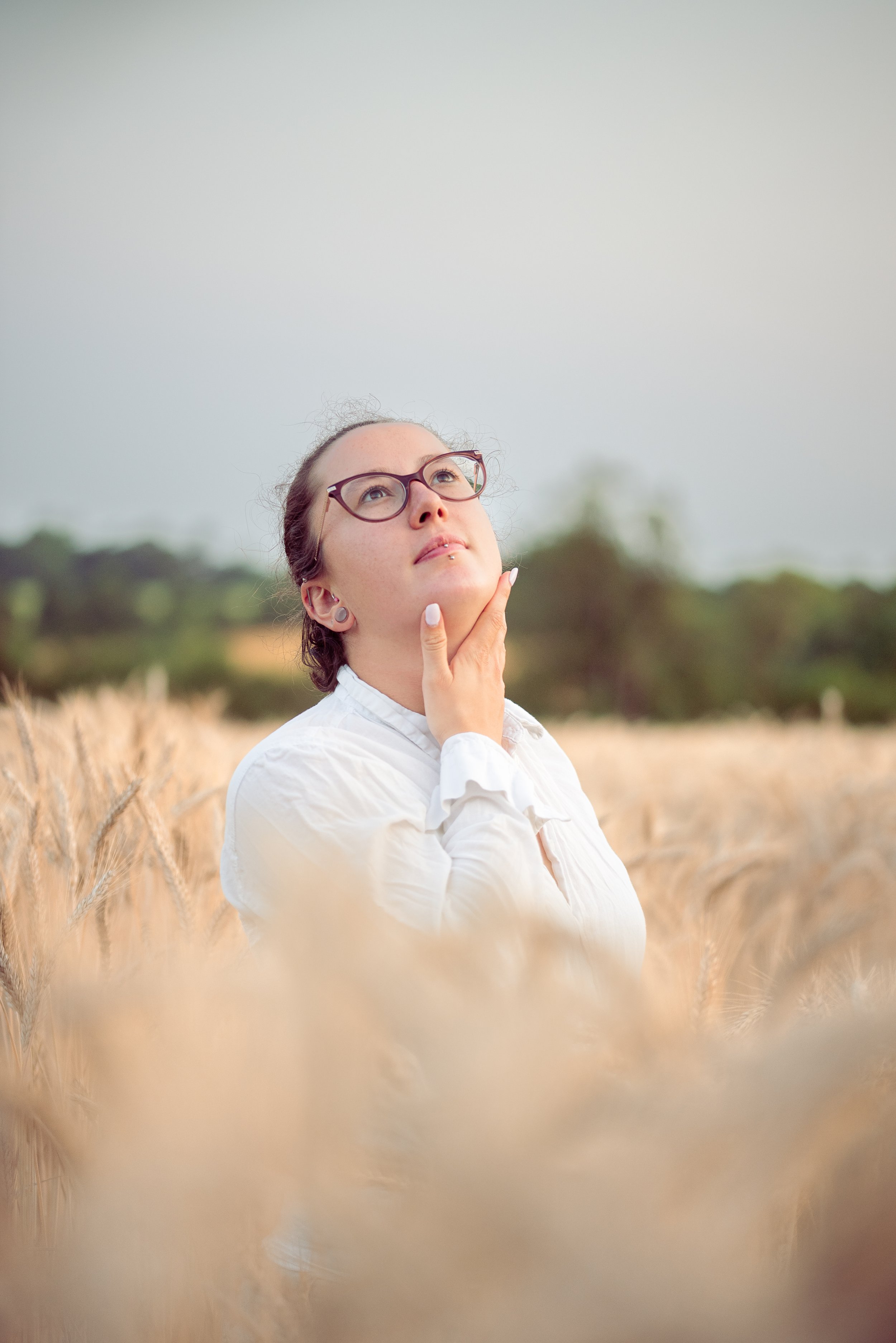 Une femme portant des lunettes et une chemise blanche se tient dans un champ de blé, regardant vers le haut avec une main sous le menton, l'arrière-plan est flou avec des arbres.