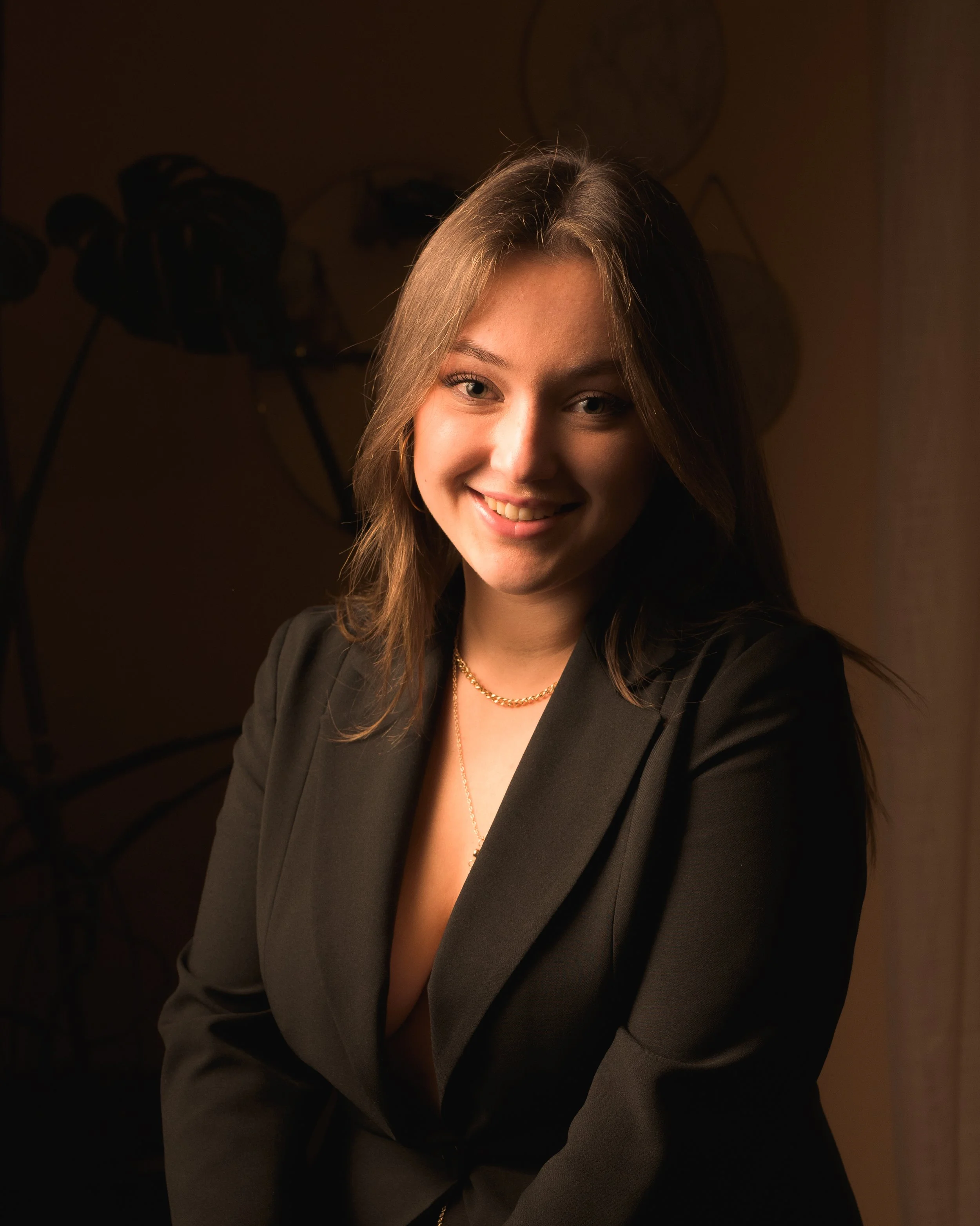 Jeune femme souriante portant un blazer noir, une chaîne et un collier, positionnée posant pour une photo en intérieur, arrière-plan sombre avec des décorations