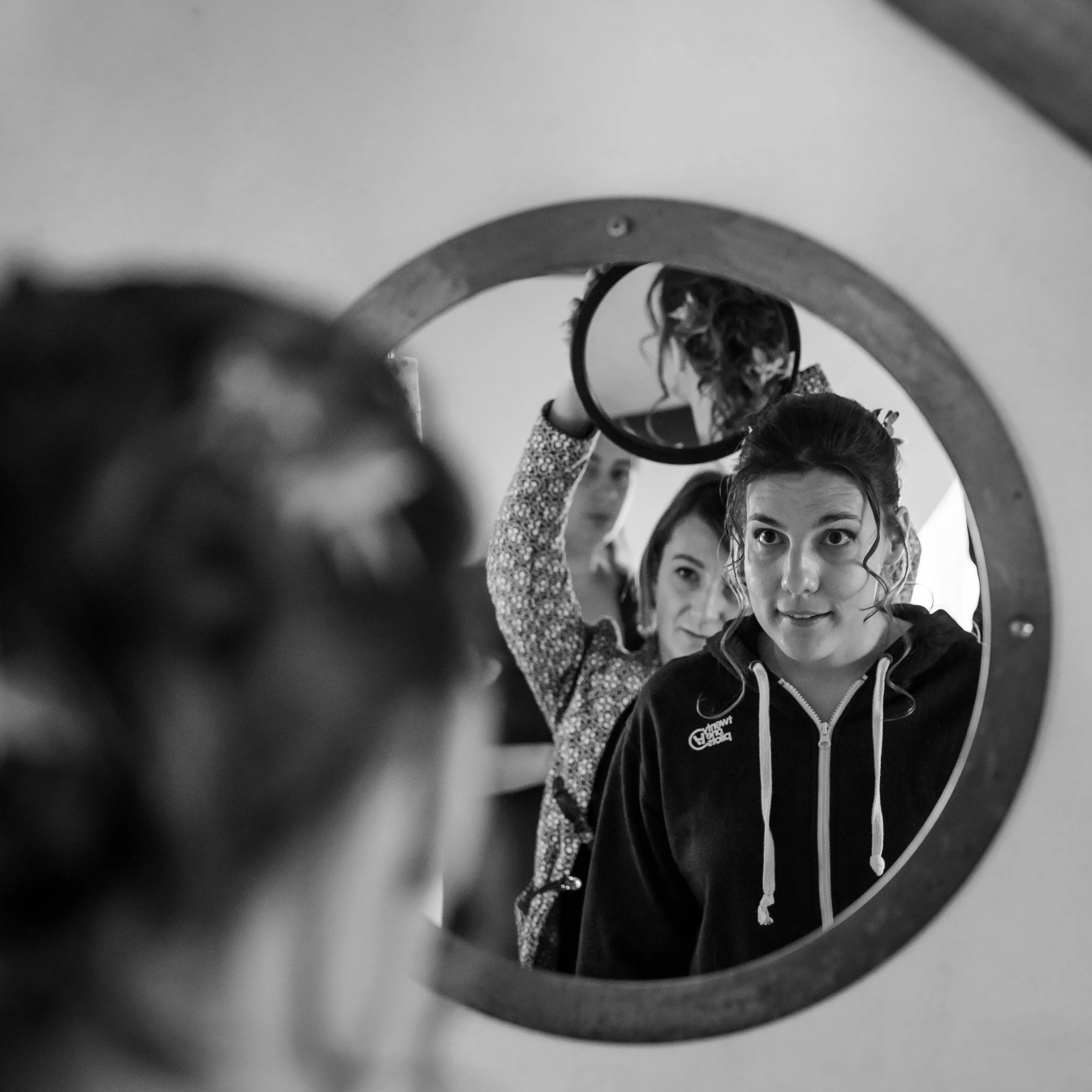 Jeune femme regardant dans un miroir, portant un sweat à capuche noir avec un logo, dans un environnement intérieur.