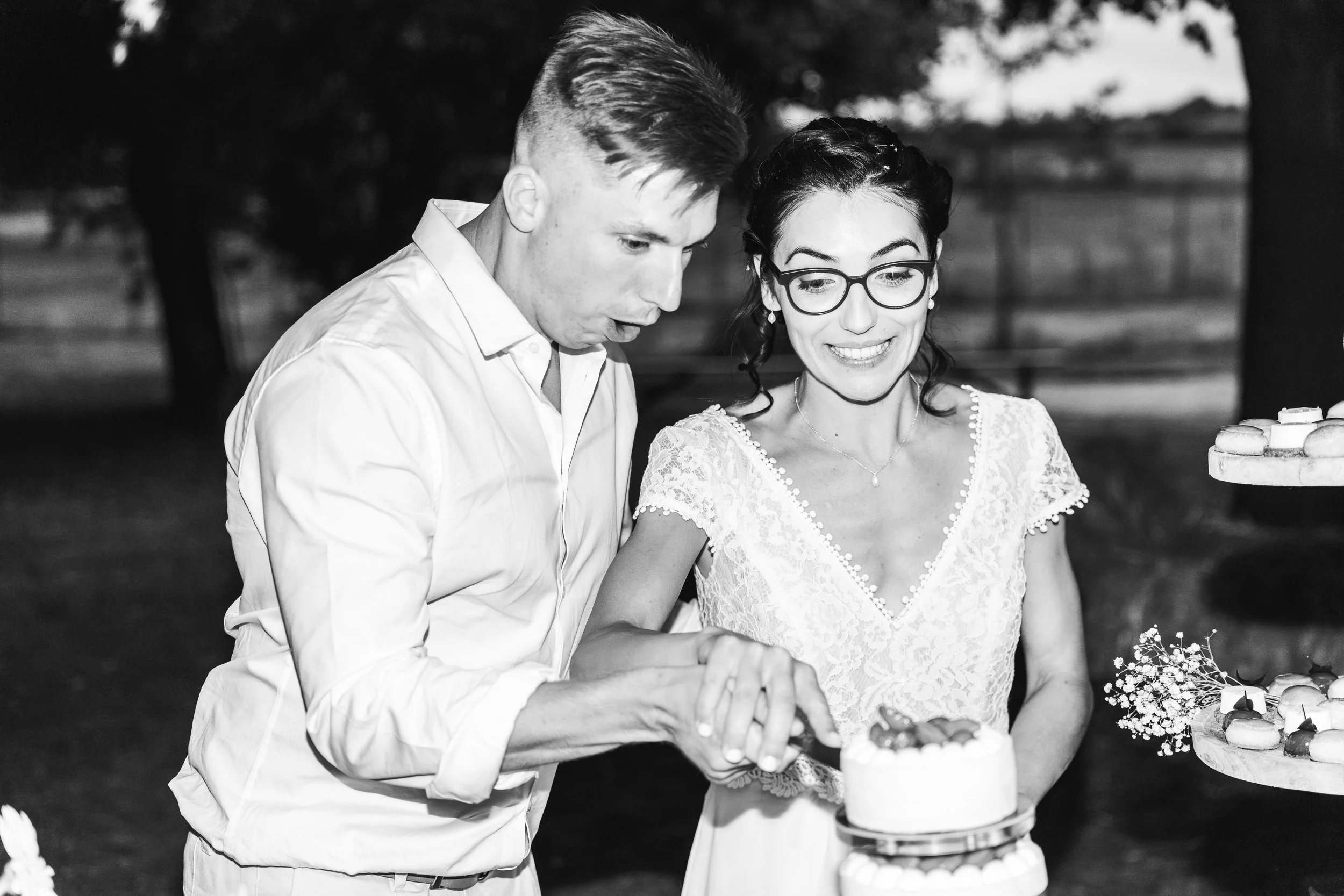 Un couple en tenue de mariage, la mariée porte des lunettes et un vêtement en dentelle, ils semblent découper un gâteau de mariage à l'extérieur. L'image est en noir et blanc.