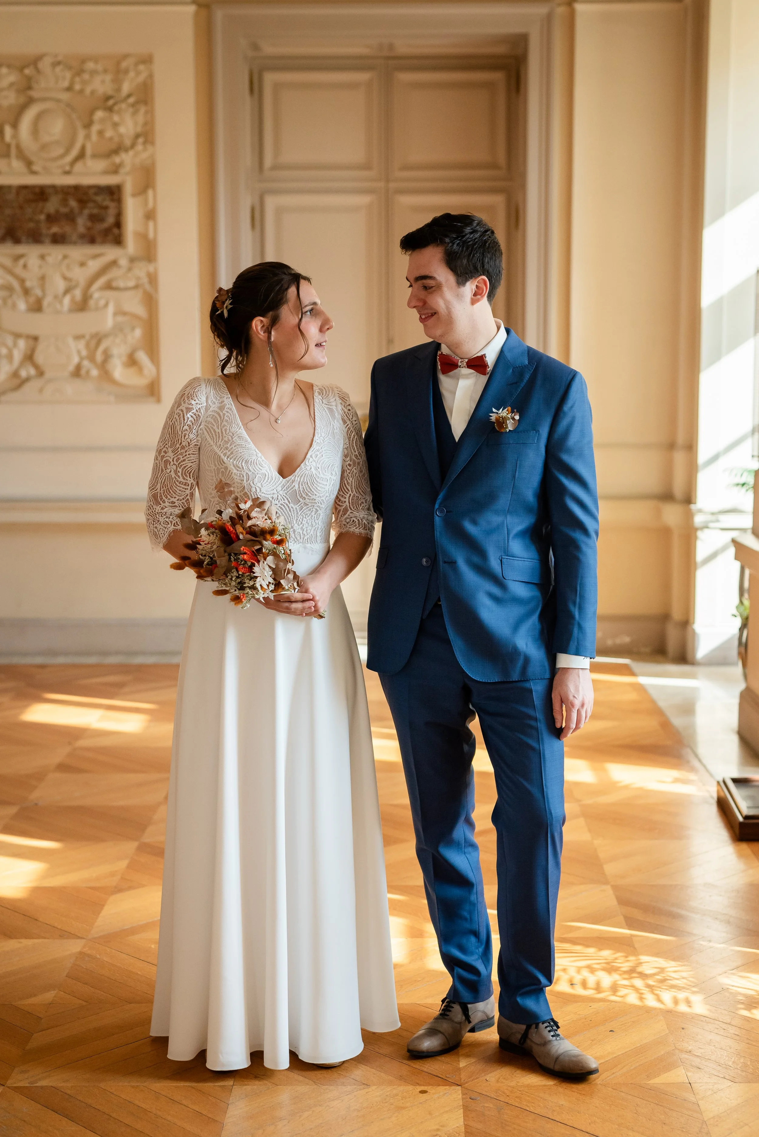 Un couple en tenue de mariage à l'intérieur d'une salle élégante avec des murs ornés et un parquet en bois. La femme porte une robe blanche avec des détails en dentelle et tient un bouquet de fleurs automnales. L'homme porte un costume bleu avec une 