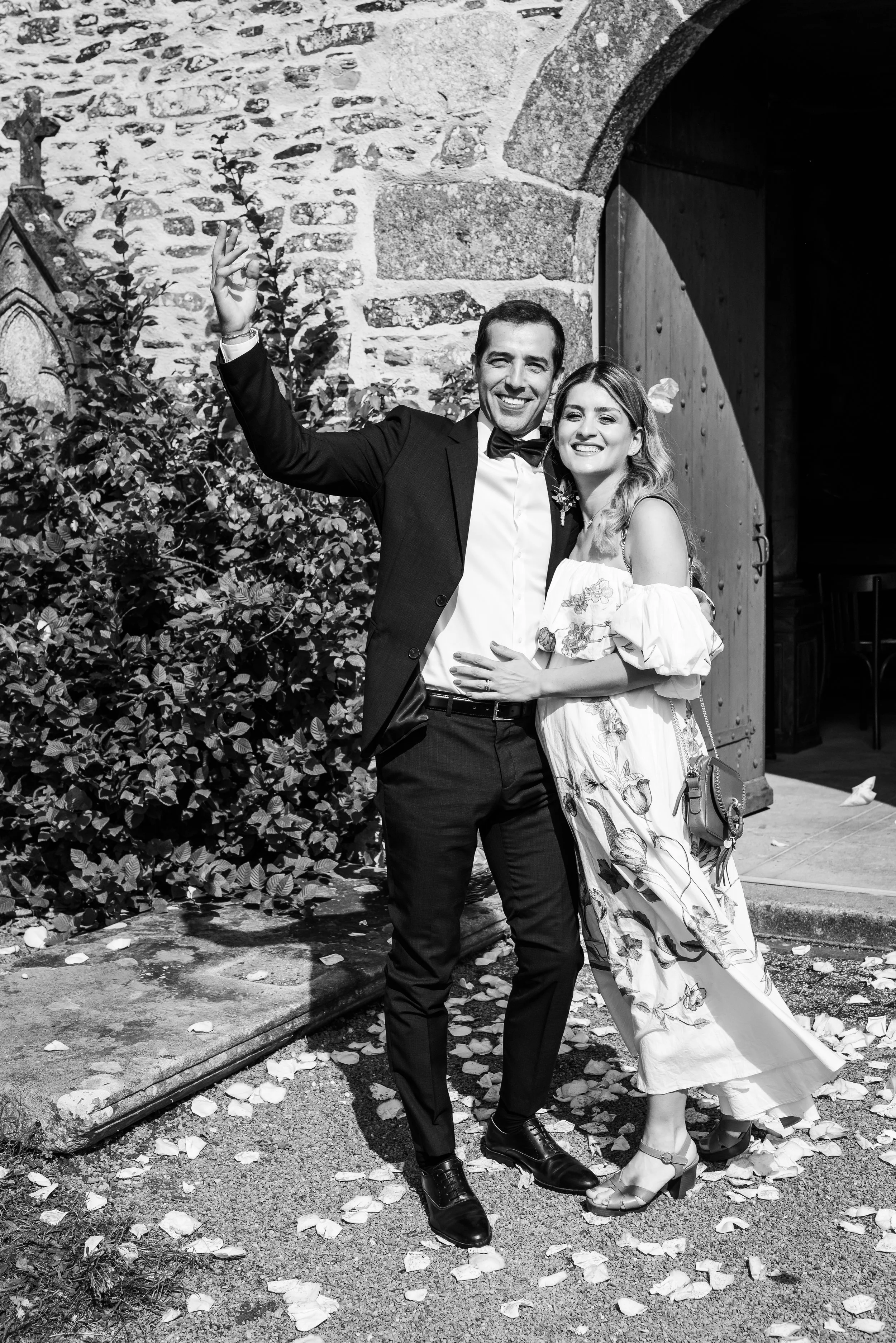 Un couple de mariés souriant, posant à l'extérieur d'une vieille église en pierre, avec des feuilles tombées au sol, en noir et blanc.