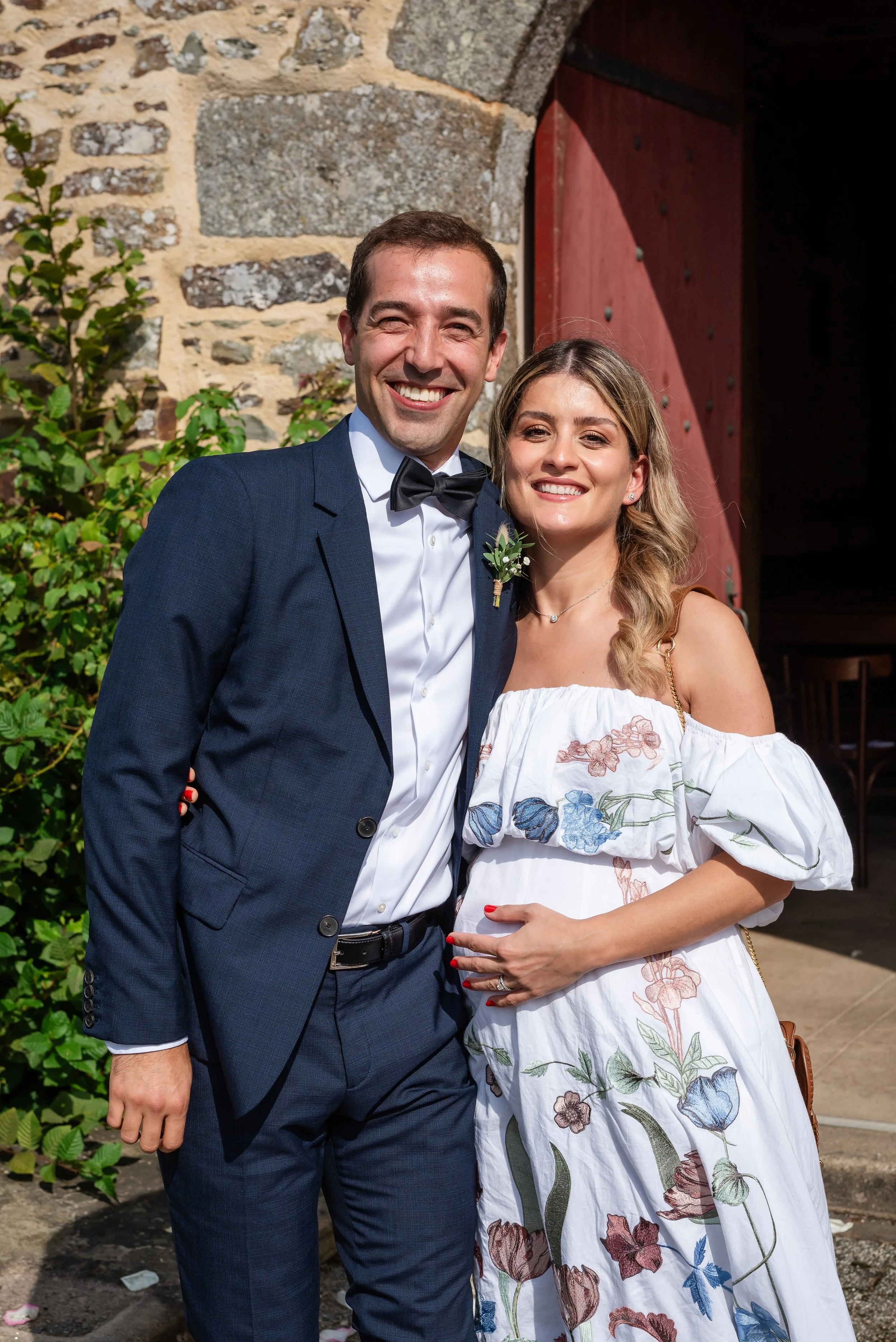 Un homme en costume de mariage et une femme en robe de mariage posent côte à côte devant un mur en pierre et une porte en bois, tous deux souriants.