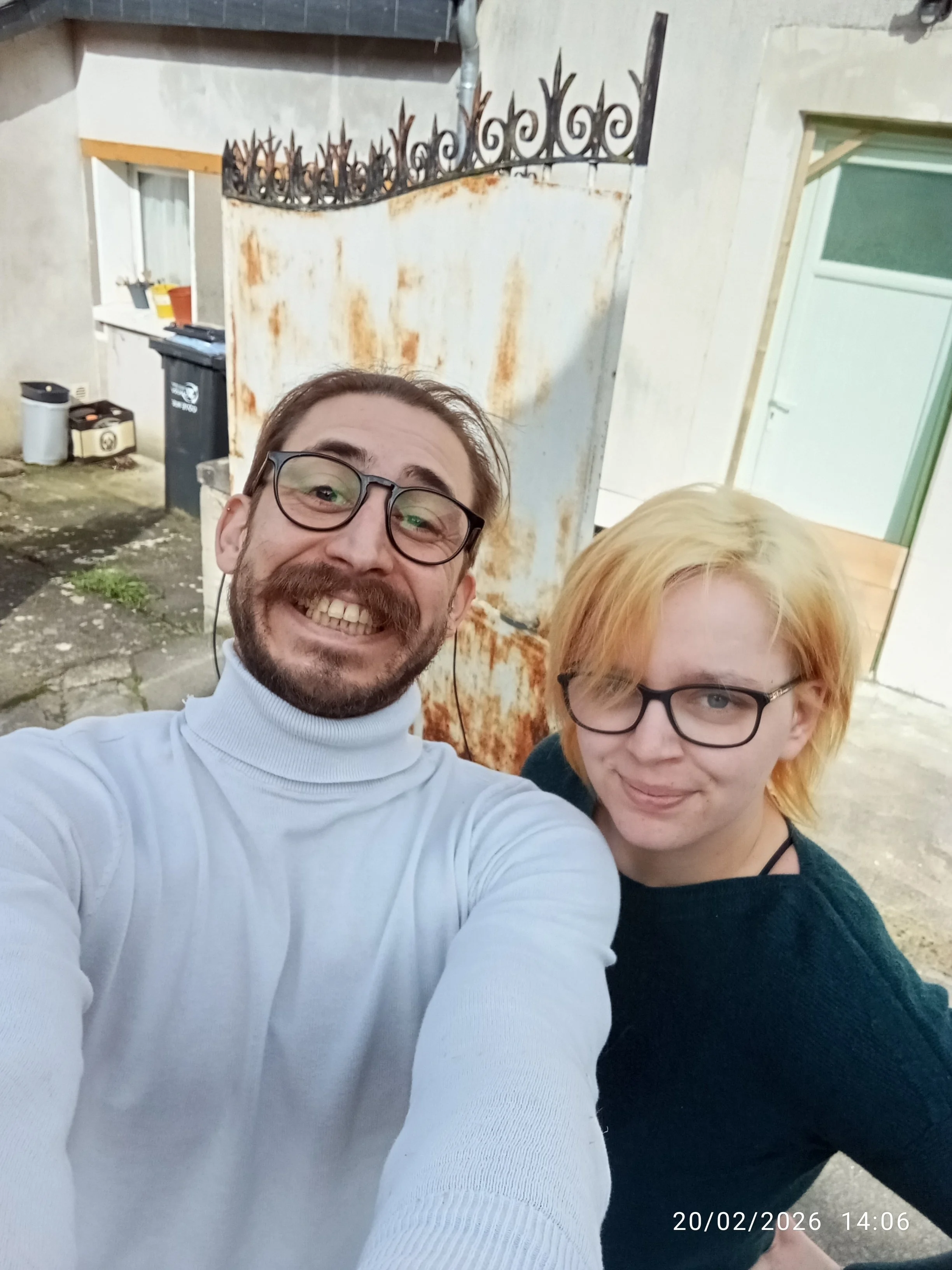 Deux personnes souriantes prenant un selfie devant une porte rouillée et un mur en béton, avec une horloge indiquant la date 20/02/2026 et l'heure 14:06.