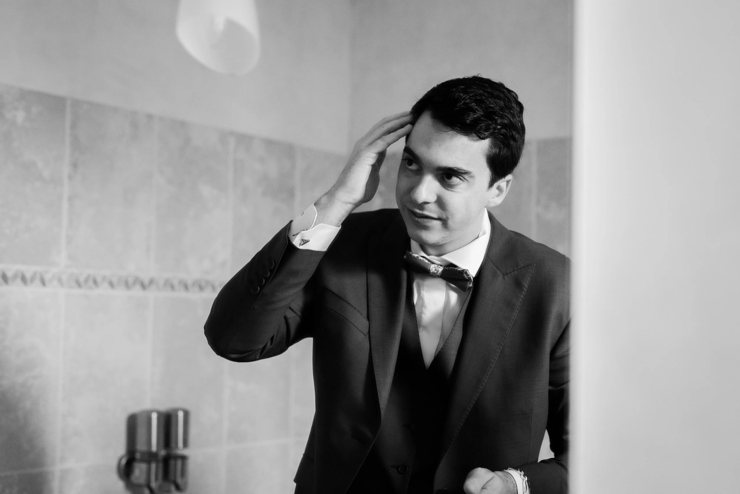 Un jeune homme en costume regardant dans un miroir, touchant sa tête, dans une pièce aux murs en céramique.