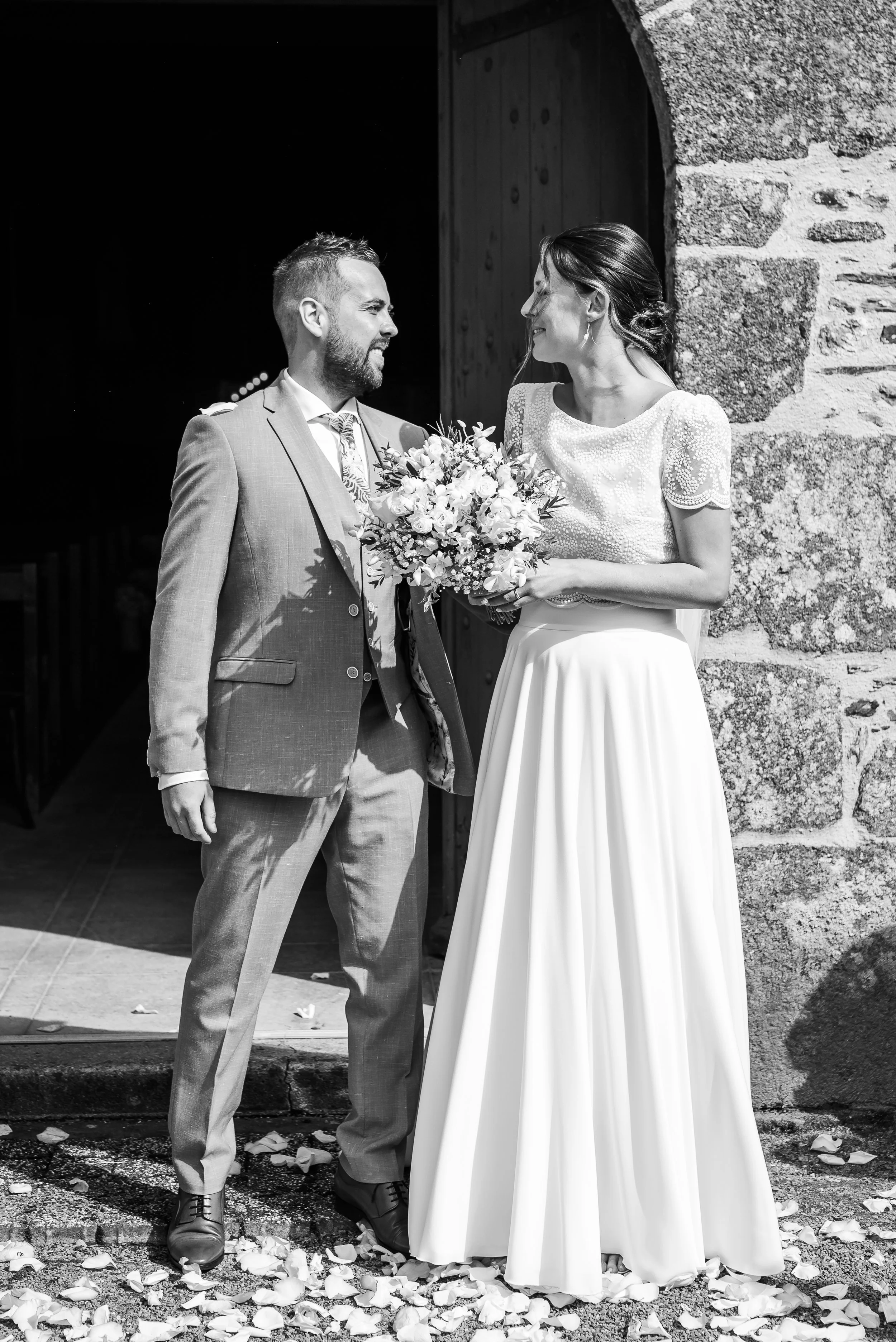 Un couple de mariés, un homme en costume et une femme en robe de mariée, échangeant un regard devant un bâtiment en pierre, la femme tenant un bouquet de fleurs blanc. La scène est en noir et blanc.