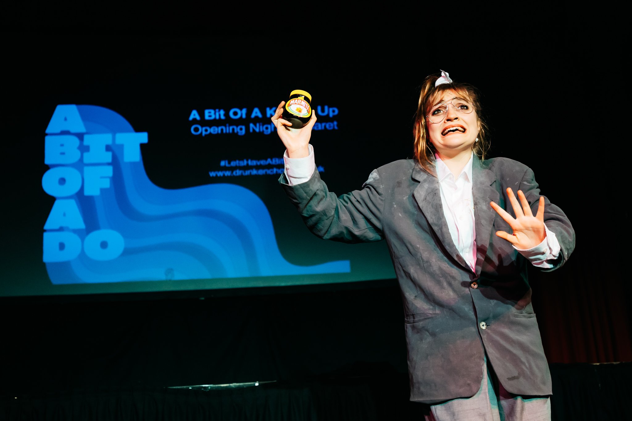  A performer stands on stage with a jar of marmite aloft in one hand and a grimace on her face. She wears a grey suit with a white shirt underneath and her long brown hair is in a pony tail. On a screen behind her are the words 'A Bit Of A Do' 