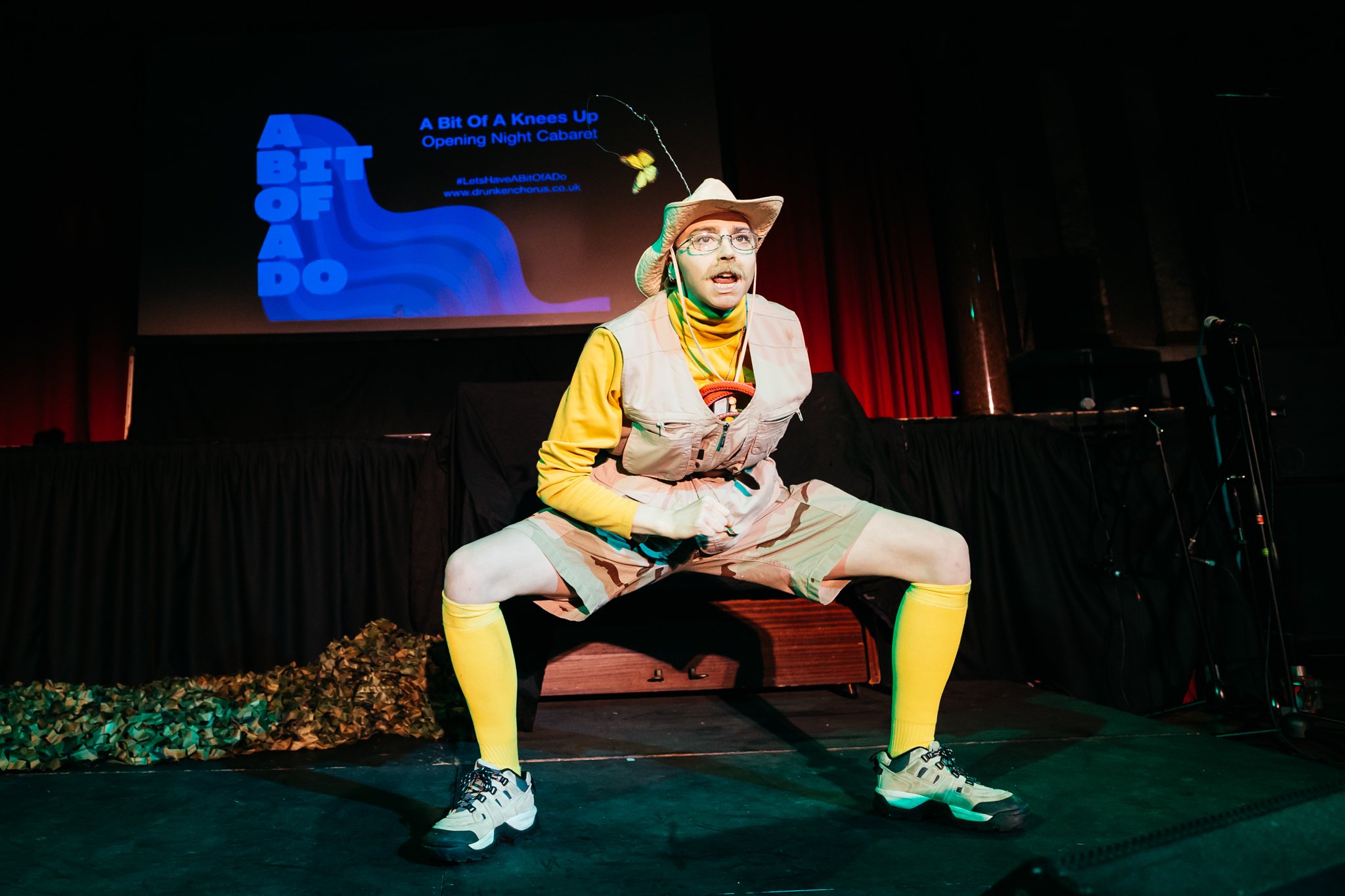  Frank, a Drag King is wearing a yellow t-shirt and knee high socks and a khaki waistcoat, shorts and explorer hat. He performs a very deep squat – he is very flexible! 