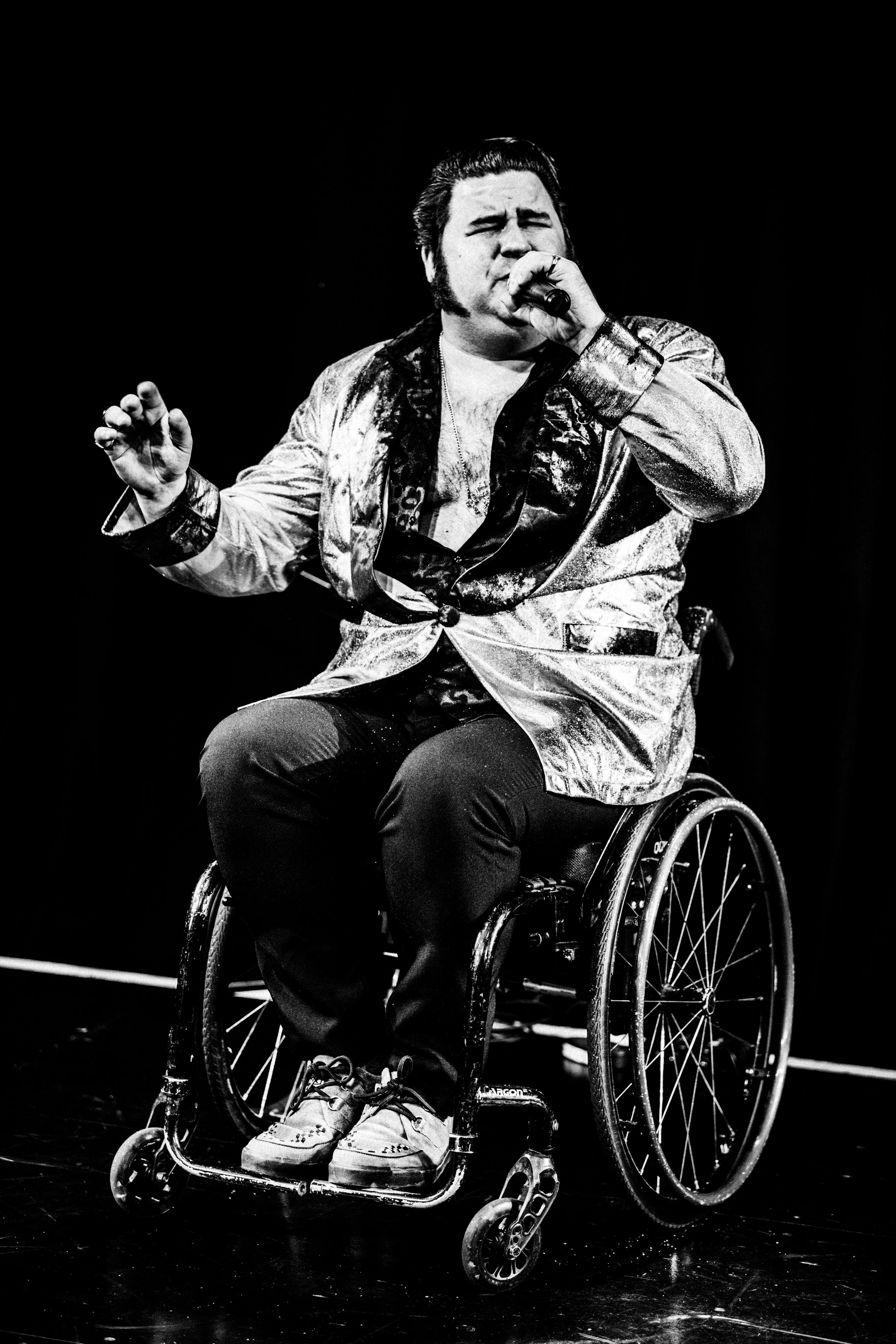 The picture is in black and white. The performer is dressed up to look like Elvis. He sings into a microphone, clearly giving his all. The performer is a wheelchair user.