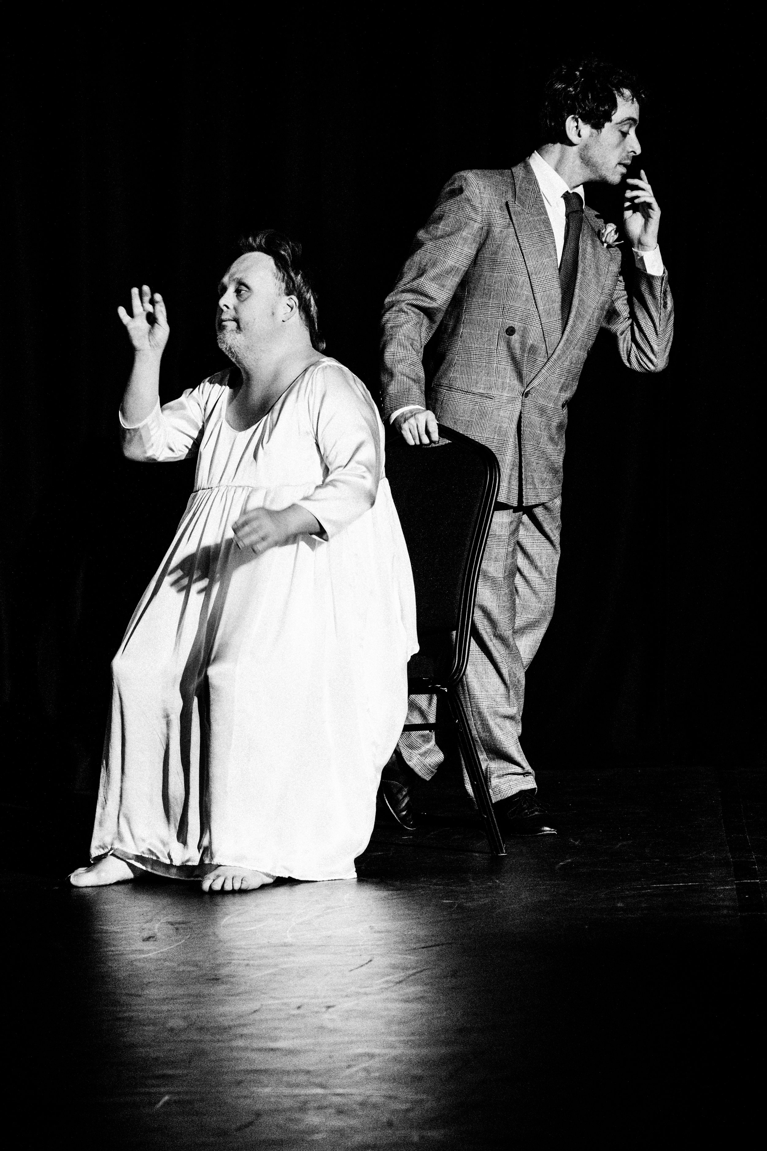 The image is in black and white and is of two performers. One sits down, looks off to the left and wears a long white dress. The other stands up, looks off to the right and wears a suit
