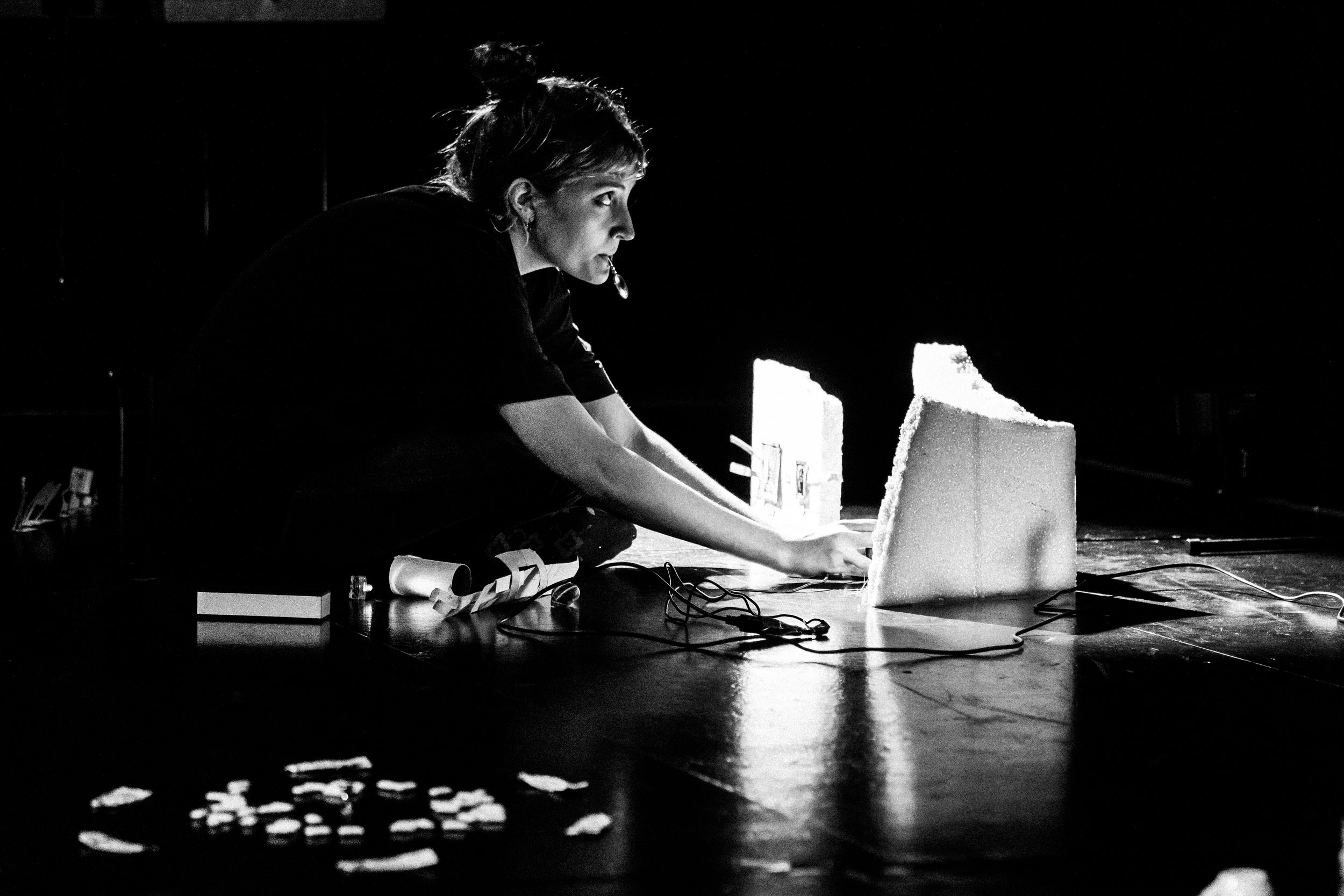 The image is black and white. The performer knees down and concentrates on something out of frame. She has a tiny magnifying glass in her mouth.