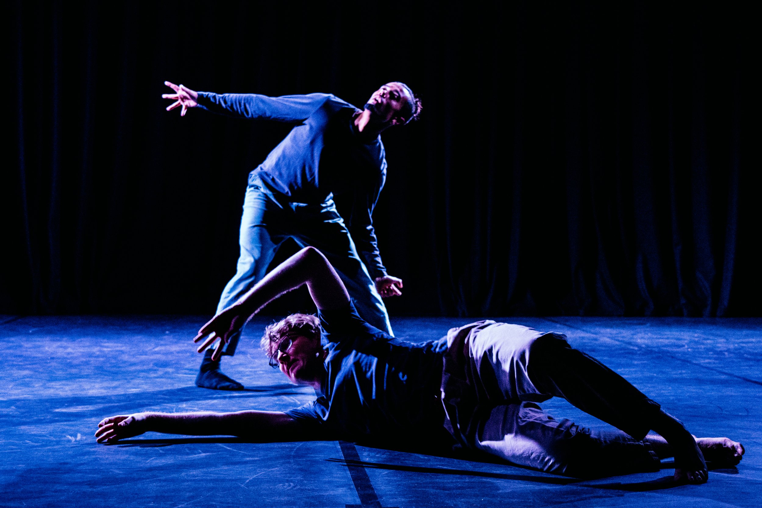 Two dancers making shapes. The stage is bathed in blue light. One dancer is on the floor the other stood up.