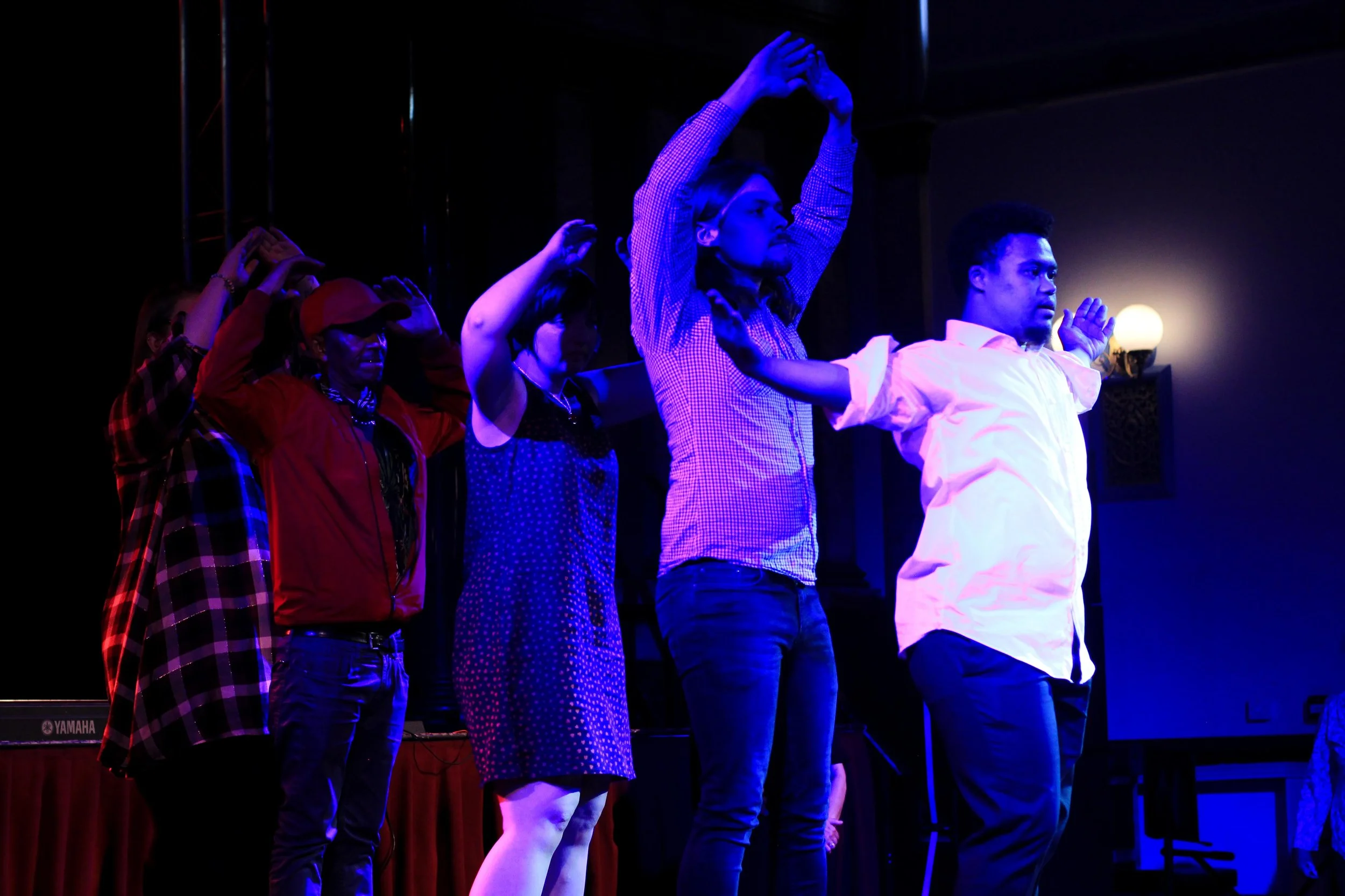The stage is bathed in blue light. 5 performers are in a line, one behind the other. The performer in front has their arms spread out wide, the performers behind all have their arms in the air