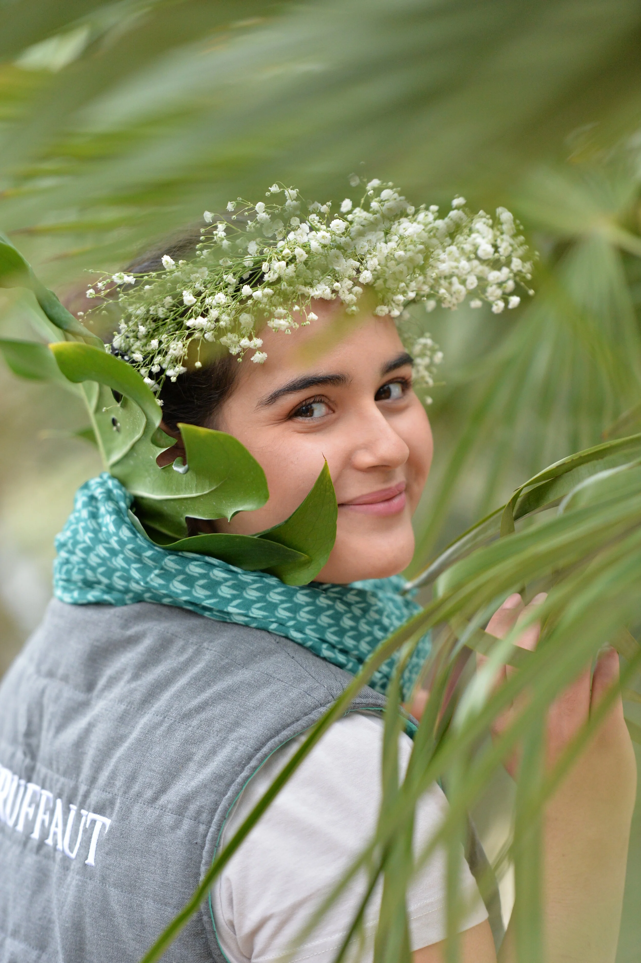 jeune femme souriante portant une couronne de petites fleurs blanches, entourée de feuilles vertes, en plein air.