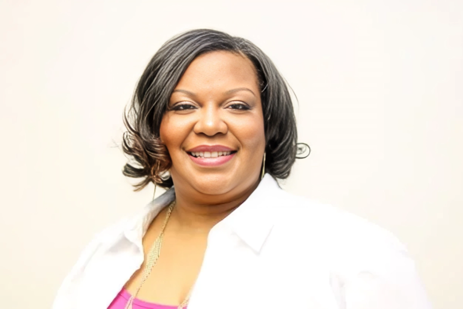 A smiling African American woman with shoulder-length dark hair, wearing a white shirt over a pink top, against a plain light background.