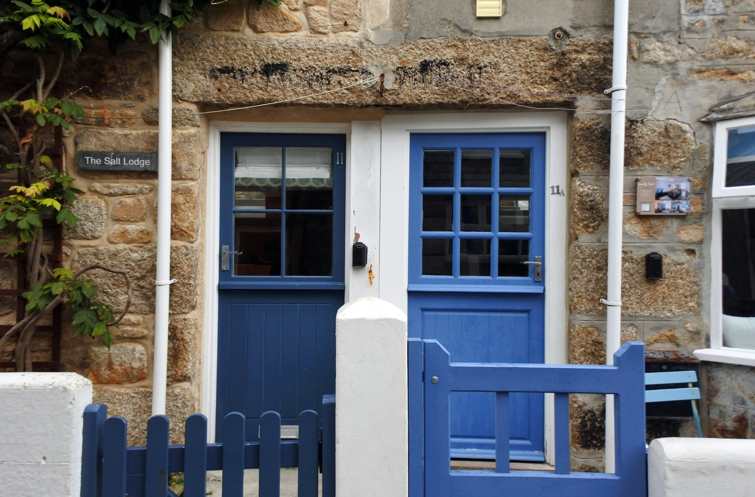 Two blue doors with white frames set in a stone wall, with a small white fence in front, a small black mailbox next to the right door, and green foliage on the left.