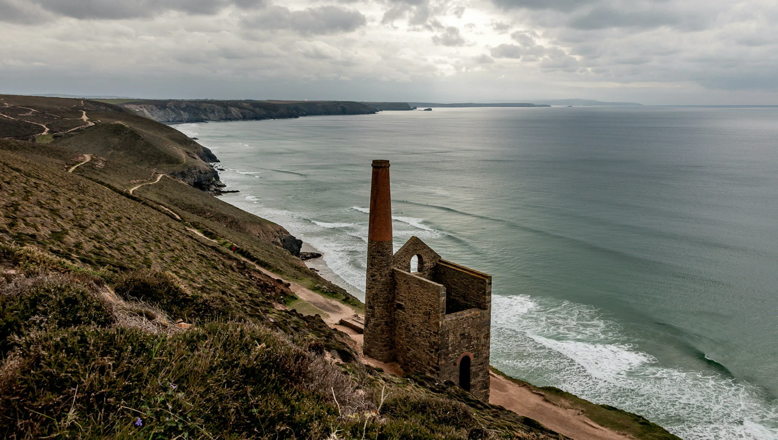 Cliffside view of the coastline with a historic stone building and a tall chimney, overlooking the ocean under cloudy skies.
