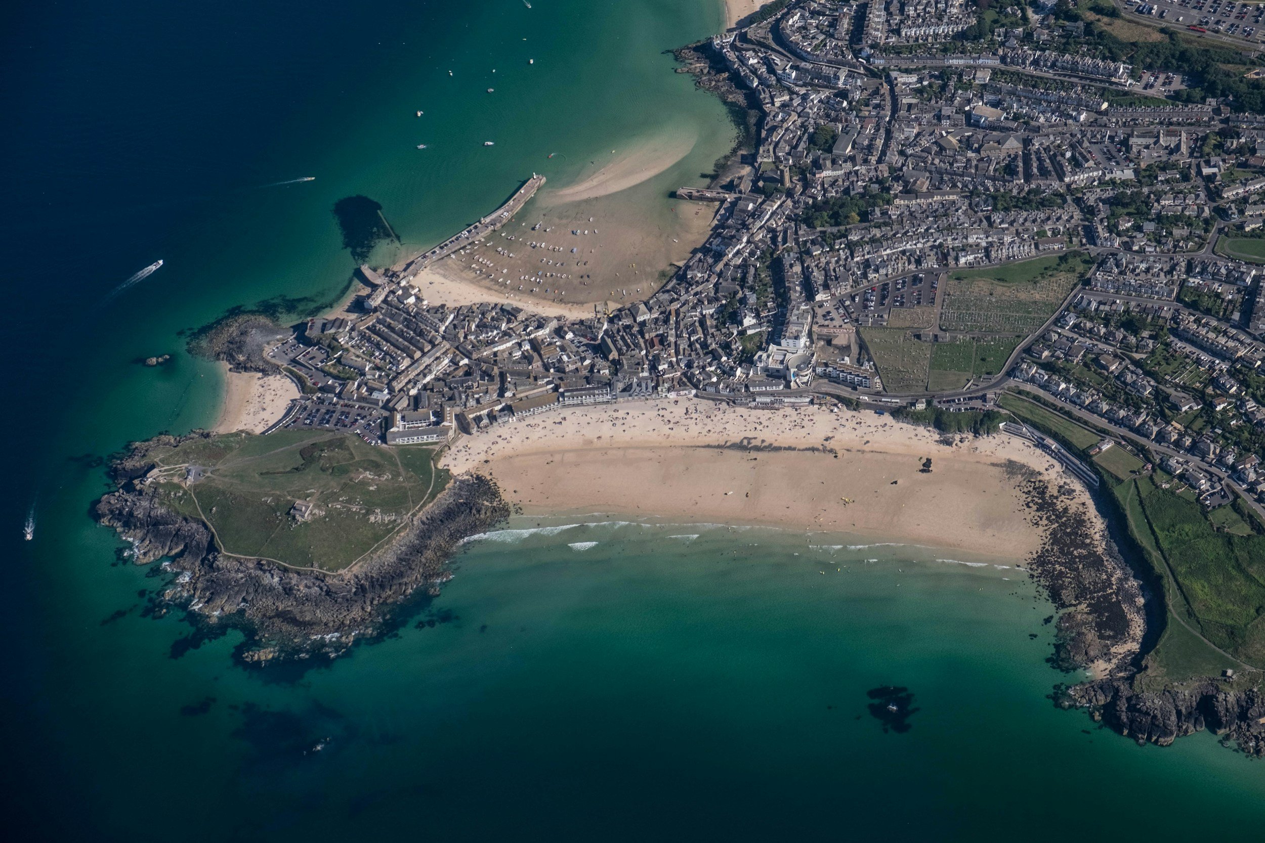 An aerial view of a small coastal town with a sandy beach, a harbor, and surrounding buildings and green areas.