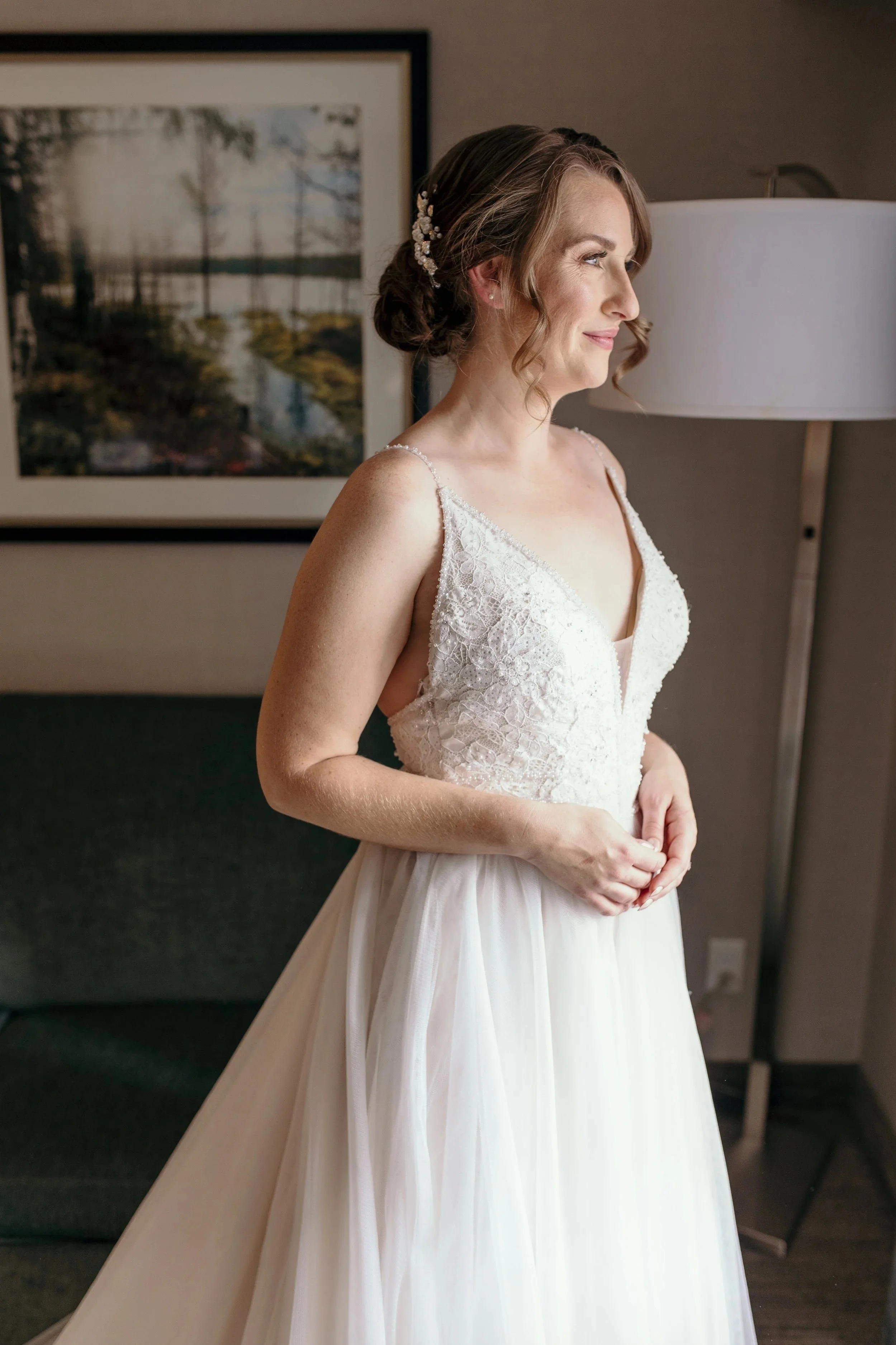 A woman in a wedding dress standing indoors near a lamp, with a framed landscape photo on the wall behind her.