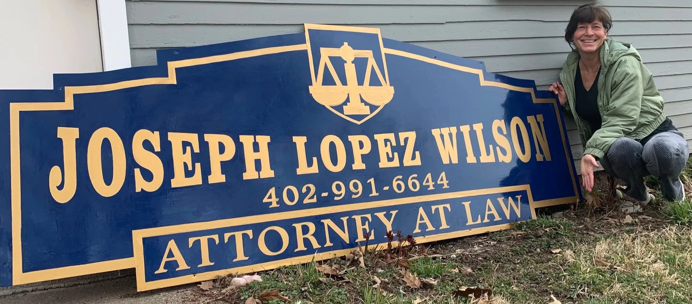 A smiling woman squatting next to a large blue and gold sign for Joseph Lopez Wilson, Attorney at Law, outside a house with light gray siding and a window.