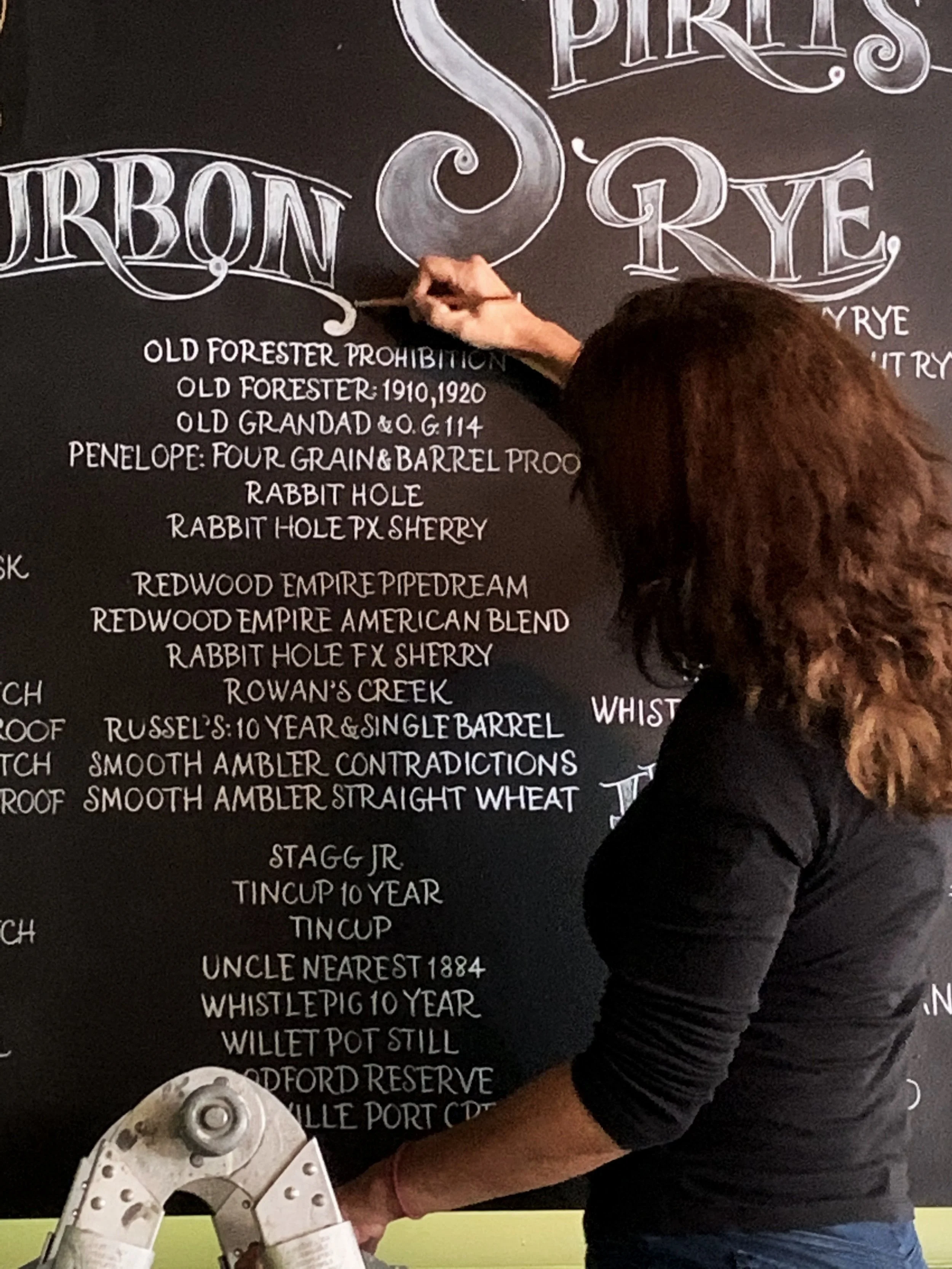 A woman writing on a black chalkboard listing different types of rye and whisky brands.