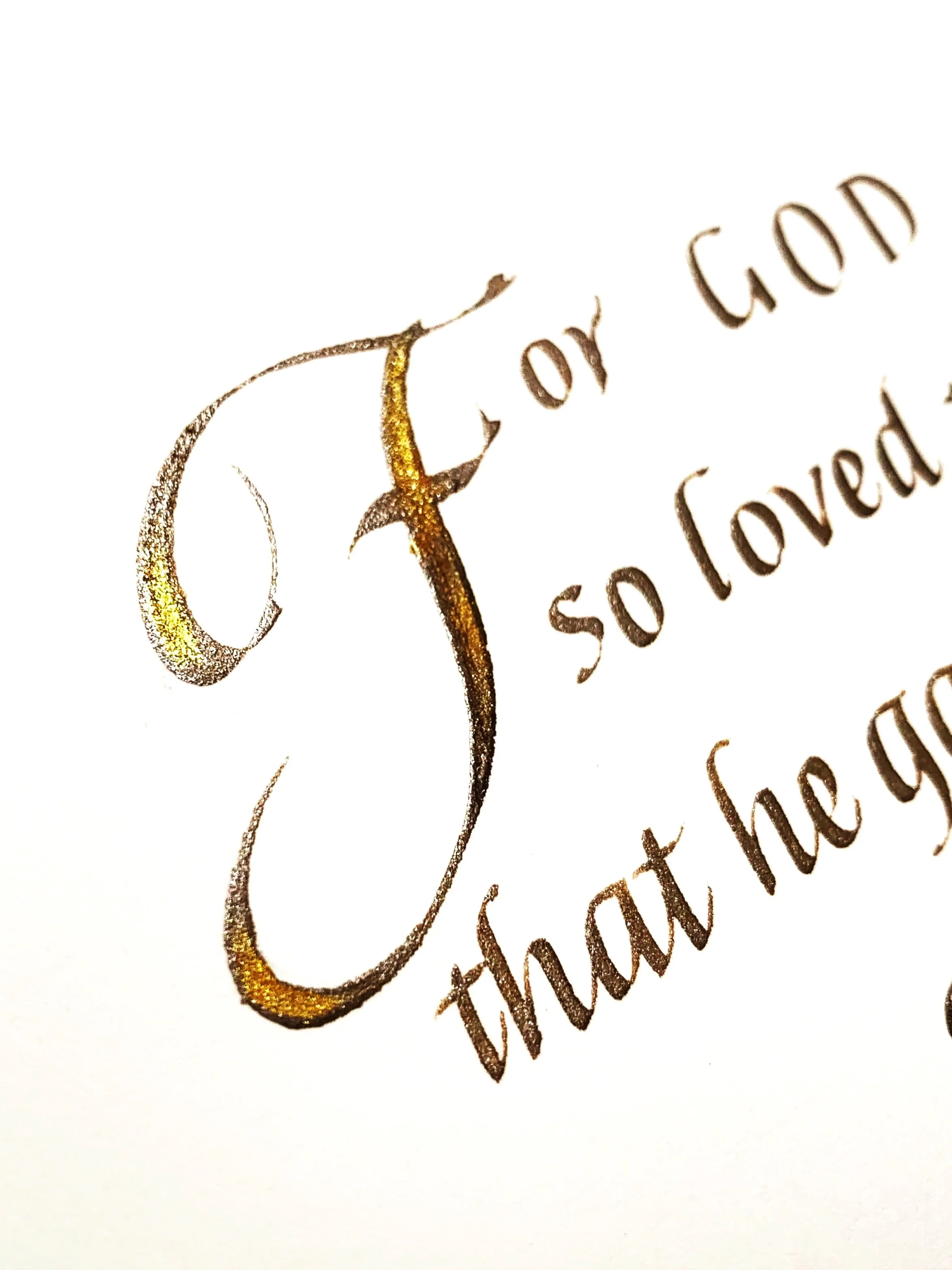 Close-up of elegant calligraphy text on white paper, featuring a large, decorative gold and black letter 'T' and words 'for God so loved'.