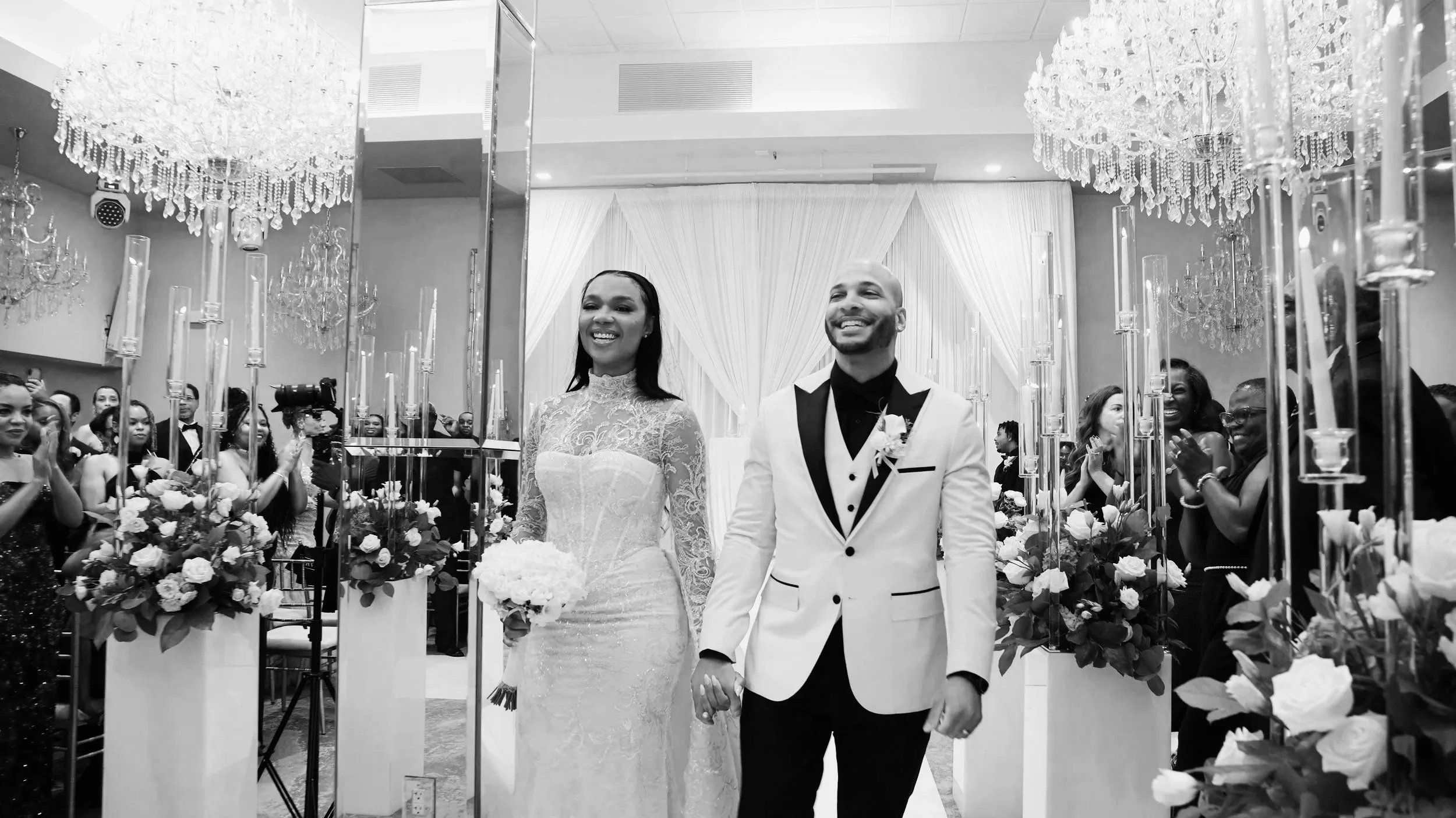 A smiling bride and groom holding hands, walking down the aisle during their wedding celebration. The bride is in a lace wedding dress, and the groom is in a tuxedo. Guests are clapping and watching in a decorated venue with chandeliers and floral arrangements.