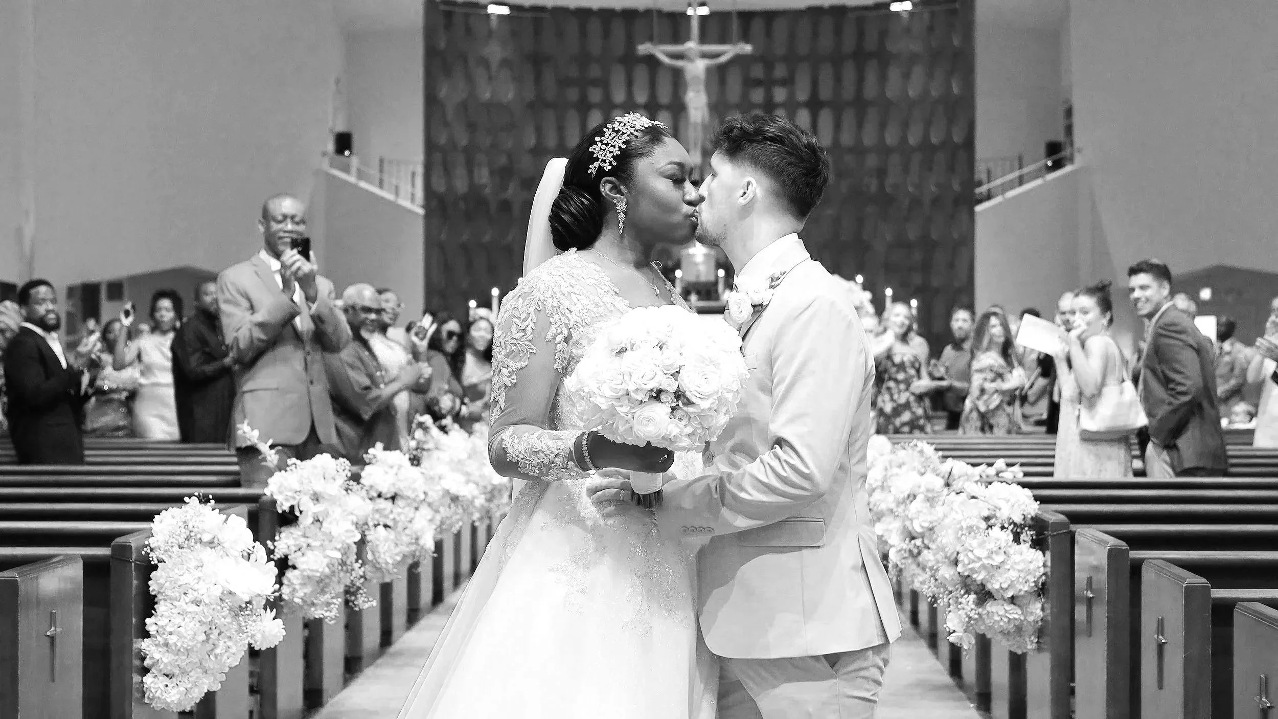 A black-and-white photograph of a wedding ceremony inside a church. The bride and groom are kissing at the altar, with the bride wearing a lace dress and veil, and the groom in a suit. The bride is holding a bouquet of flowers. Guests are standing on both sides of the aisle, some taking photos and smiling.