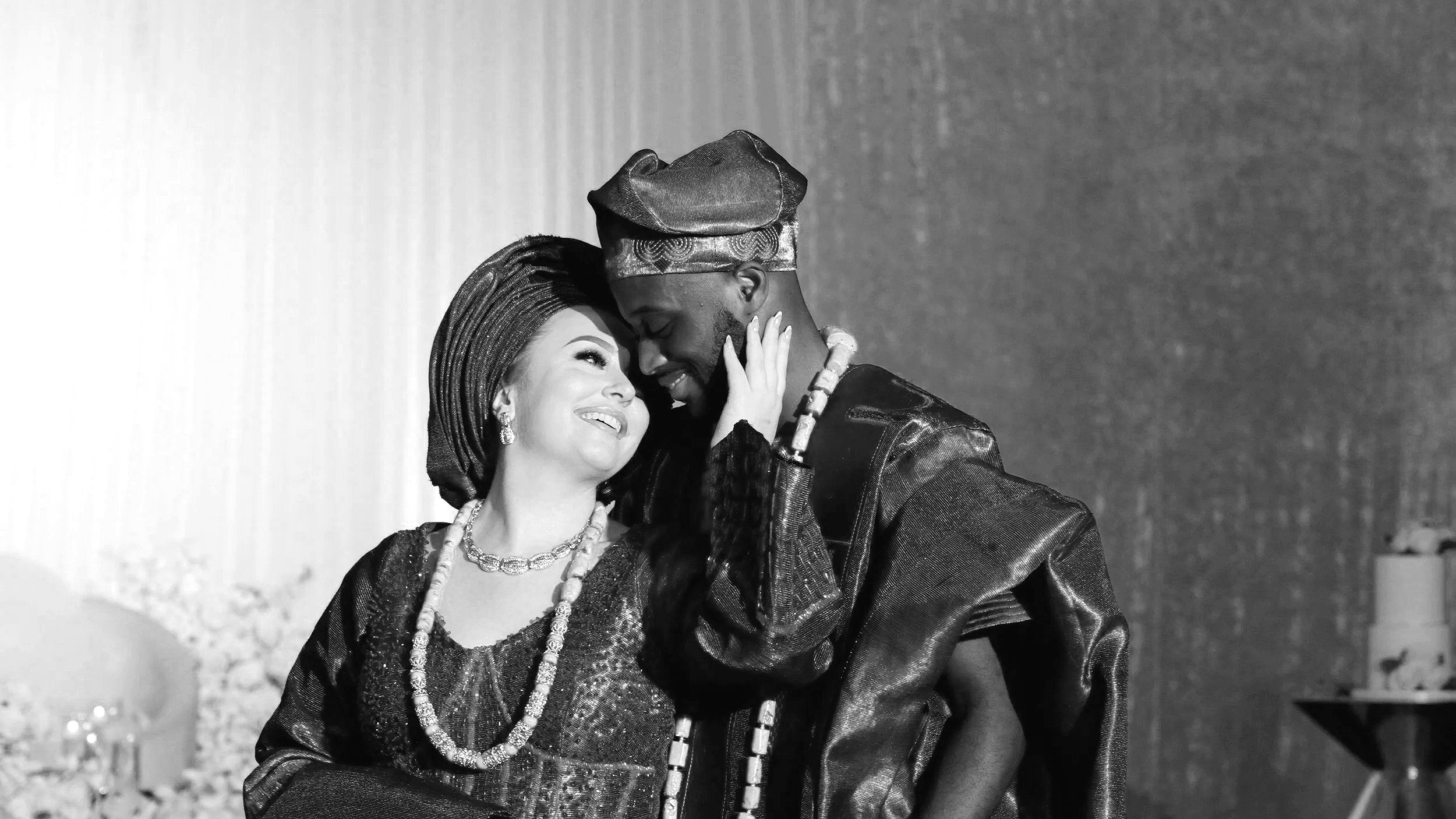 A woman and man, both dressed in traditional African attire, are smiling and embracing each other at a celebration or event.