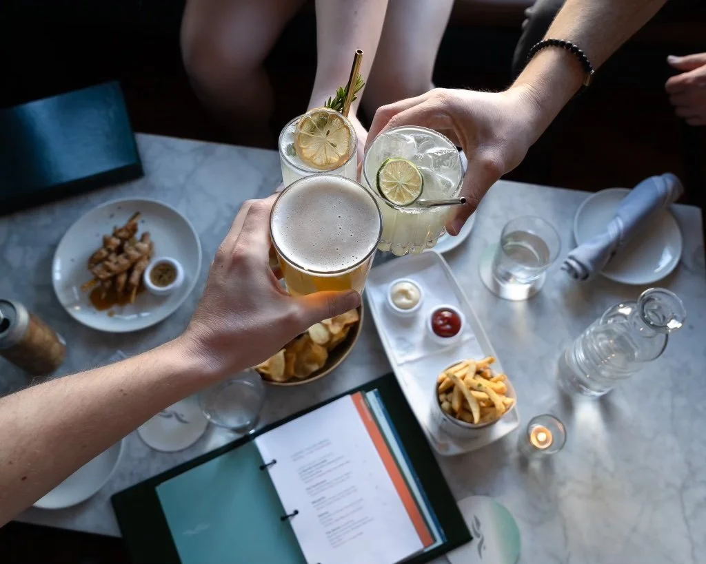 Three people toasting with drinks, including beer and cocktails with lemon and lime slices, at a table with appetizers, a candle, and a menu.