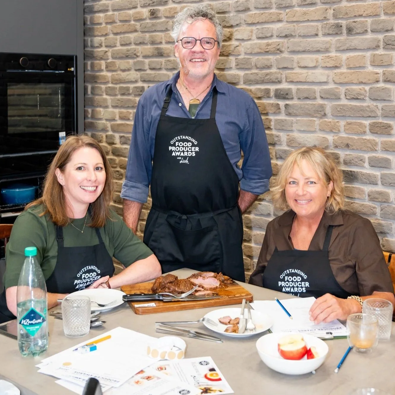 Three people at a food event wearing aprons labeled 'Outstanding Food Producer Awards.' The man in the middle is standing, and two women are seated at a table with a tray of sliced meat, plates, utensils, a water bottle, a bowl of apple slices, and papers.