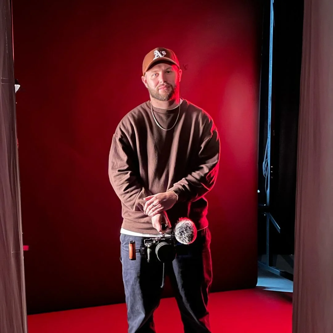 A man standing in front of a red and black background, holding a camera with a microphone attached, wearing a brown sweatshirt, black pants, a cap, and a chain necklace.
